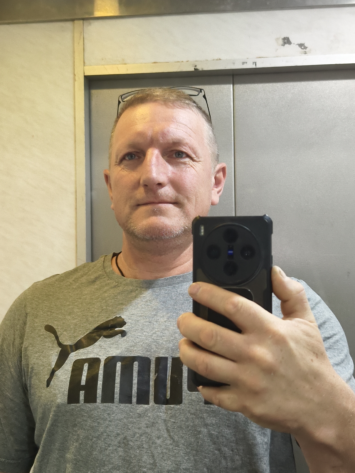 A middle-aged man is taking a selfie in what appears to be an elevator. He has short, light-colored hair, blue eyes, and a slightly stubbled face. He is wearing glasses perched on his head and a grey t-shirt with a Puma logo. He's holding a black smartphone with a prominent circular camera module on the back. The elevator's interior consists of a light-colored panel on one side and a stainless-steel door on the other. The location cannot be forensically confirmed as Petah Tikva, Israel without additional information or corroborating evidence in the image.