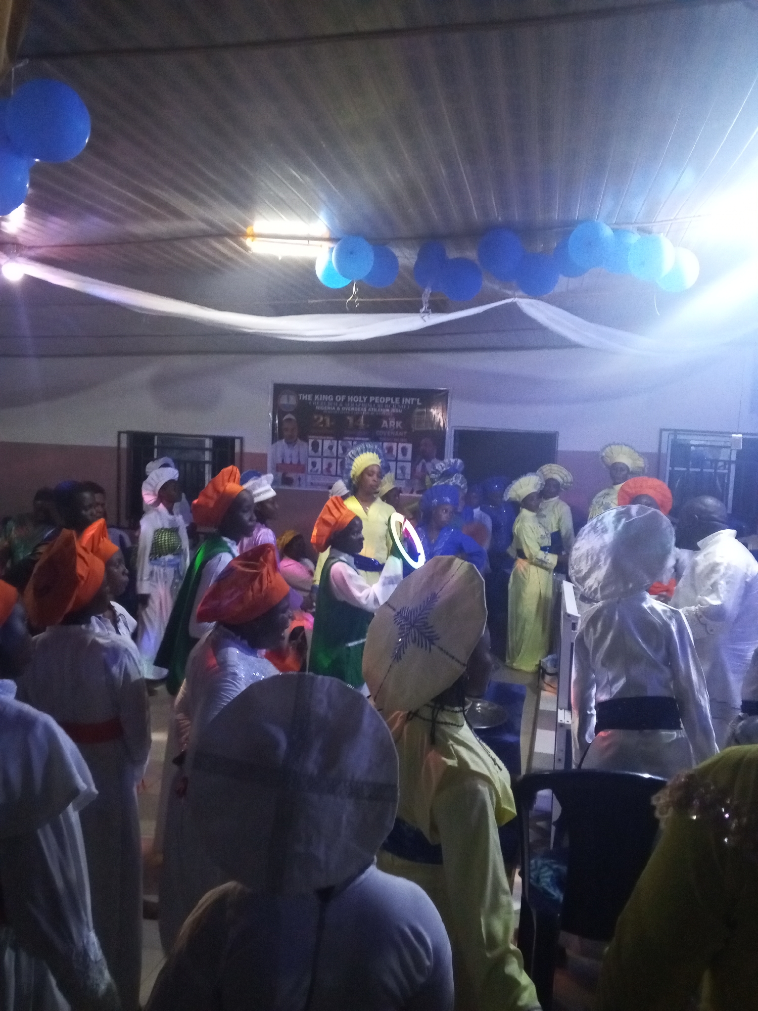 A group of people, presumably members of a religious organization, are gathered indoors in what appears to be a hall decorated with blue balloons and white fabric. The location is identified as Lokoja, Nigeria. The attendees are dressed in distinct attire, including white gowns, colorful head coverings (orange, yellow, white, blue), and some with belts. A banner with images of a man and text related to "THE KING OF HOLY PEOPLE INT'L" is visible in the background. Interactions include people standing and appearing to engage in a ceremony or event. The environment is dimly lit.