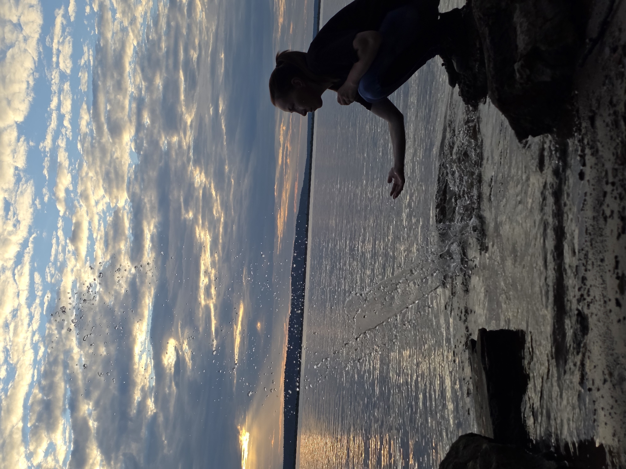 The image depicts a silhouette of a person, likely a woman, crouched on the rocky shore of a large body of water, possibly a river or lake, with a picturesque sunset in the background. The sky is filled with dramatic clouds, tinged with warm sunset hues of orange and yellow, interspersed with shades of blue and grey. The water reflects the sky's colors, creating a shimmering surface.
The person is wearing dark clothing and is captured in the act of splashing water with their hand, creating a spray of droplets that are illuminated against the cloudy sky. Their posture suggests a moment of playfulness or contemplation. The foreground shows the dark, textured rocks and sand of the shoreline, with some water lapping at the edge. The distant shore is visible as a dark, undulating line against the horizon.
The image appears to have been taken at dusk, with the low angle of the sun creating long shadows and enhancing the dramatic effect of the clouds. The overall mood is serene and evocative, capturing a moment of connection with nature.
This scene is consistent with the natural landscapes found in the vicinity of the city of Chaikovsky, Russia, which is situated on the Kama River. The presence of a wide river and the natural topography of the shoreline are characteristic of this region.