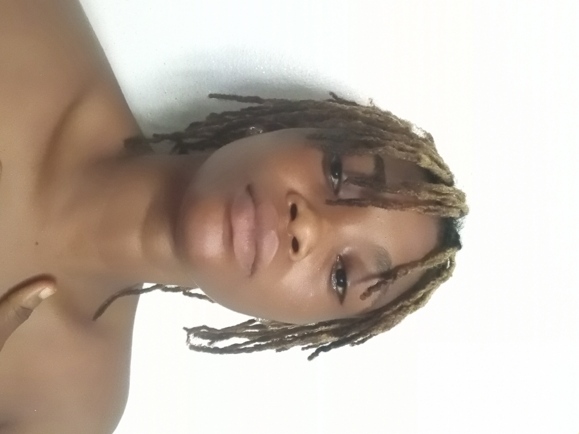 A portrait of a dark-skinned person with shoulder-length blonde dreadlocks, seemingly indoors with a plain white background. The person is facing the camera with a neutral expression, and a part of their shoulder is visible. There is no indication that the scene is located in Shagamu, Nigeria.