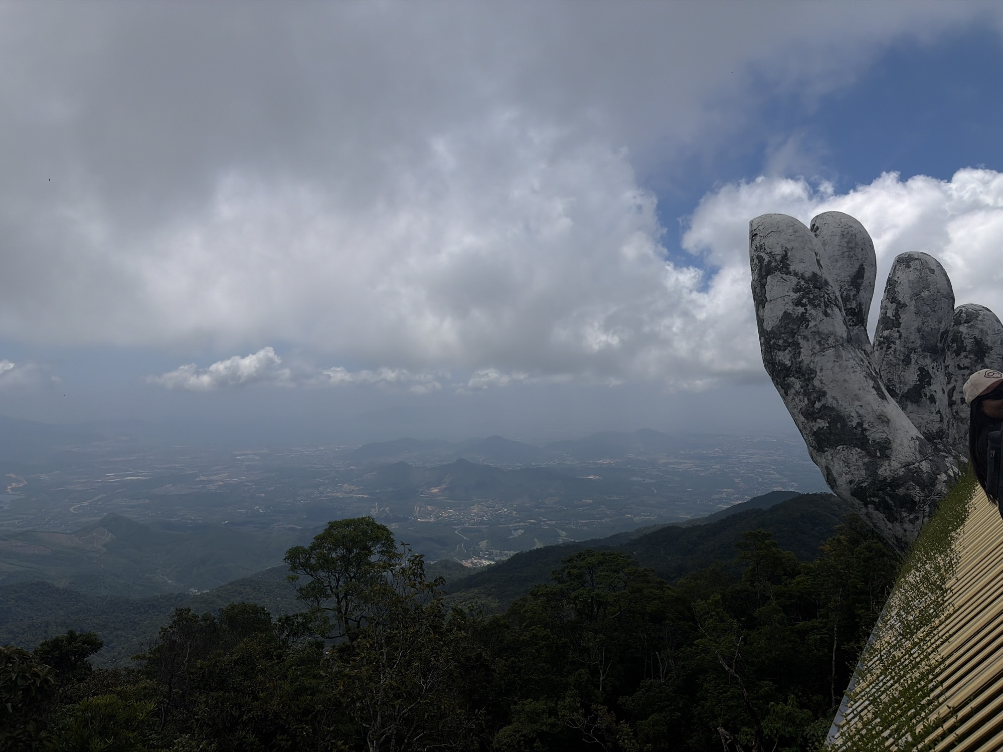 The image depicts a scene from the Golden Bridge (Cầu Vàng) in the Bà Nà Hills near Đà Nẵng, Vietnam. The foreground features a view of the landscape covered in dense green forest and distant valley and hills covered with haze. In the immediate foreground are the iconic giant stone hands holding up the pedestrian bridge. The sky is filled with thick gray clouds mixed with patches of blue sky. A small section of the bridge railing is also visible. A person partially enters the frame on the right.