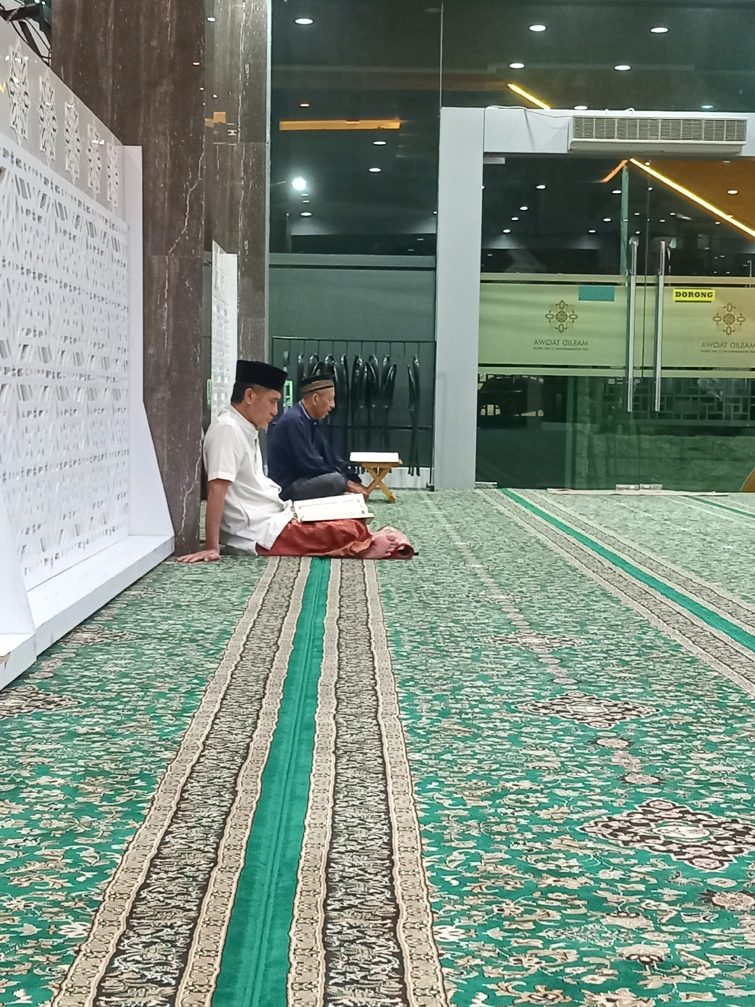 In the city of Randuagung, Indonesia, two men are seated on a green carpet with an intricate pattern in a prayer room. The man in the foreground, wearing a white shirt, dark pants, and a black peci (cap), is sitting with his left hand resting on the carpet. He has a book open on his lap, with a red cloth beneath it. The second man, in the background, wears a dark blue shirt and a peci. He is seated on a small wooden stool. Both men are facing the same direction, suggesting they are engaged in prayer or religious study. The room features a modern aesthetic, with large glass windows, a reflective ceiling with overhead lights, and a decorative white panel on the left wall. The carpet extends throughout the visible area, with a prominent green stripe running down its center. The overall impression is one of a serene and contemporary prayer space.