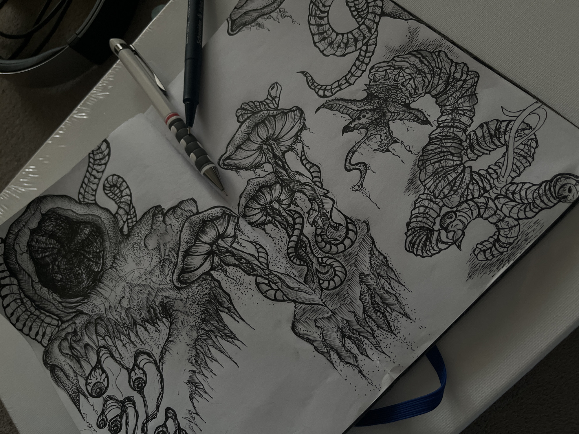 The media file presents an overhead, angled view of two open sketchbooks or sheets of paper displaying intricate, dark-themed pen and ink drawings. Two writing instruments, a white/silver mechanical pencil/pen and a black fineliner pen (partially visible with "Schneider" text), rest across the artwork, suggesting recent creative activity.
The drawings feature surreal and fantastical organic forms. On the left sheet, a large, complex illustration depicts a monstrous or alien entity, characterized by a prominent dark, textured orb or "eye," jagged protrusions, and several smaller, stalked eye-like structures. This piece employs extensive stippling, hatching, and cross-hatching to create depth and texture.
The right-hand pages showcase additional sketches. One section illustrates multiple mushroom-like forms with detailed gills and caps, some intertwined with serpentine or tentacle-like elements. Another area features elongated, creature-like figures: one is a serpentine body with a reptilian or demonic head, complete with sharp features and small wings, its body covered in textured, wrapped patterns; a simpler coiled, textured snake-like form is also visible.
The scene is set indoors, likely on a desk or table, with dim lighting that creates a shadowy ambiance. The background is dark and out of focus, offering no specific details. The artwork's style evokes a sense of macabre, dark fantasy, or horror. There are no visible cues within the image itself to indicate the location context of Sofia, Bulgaria.