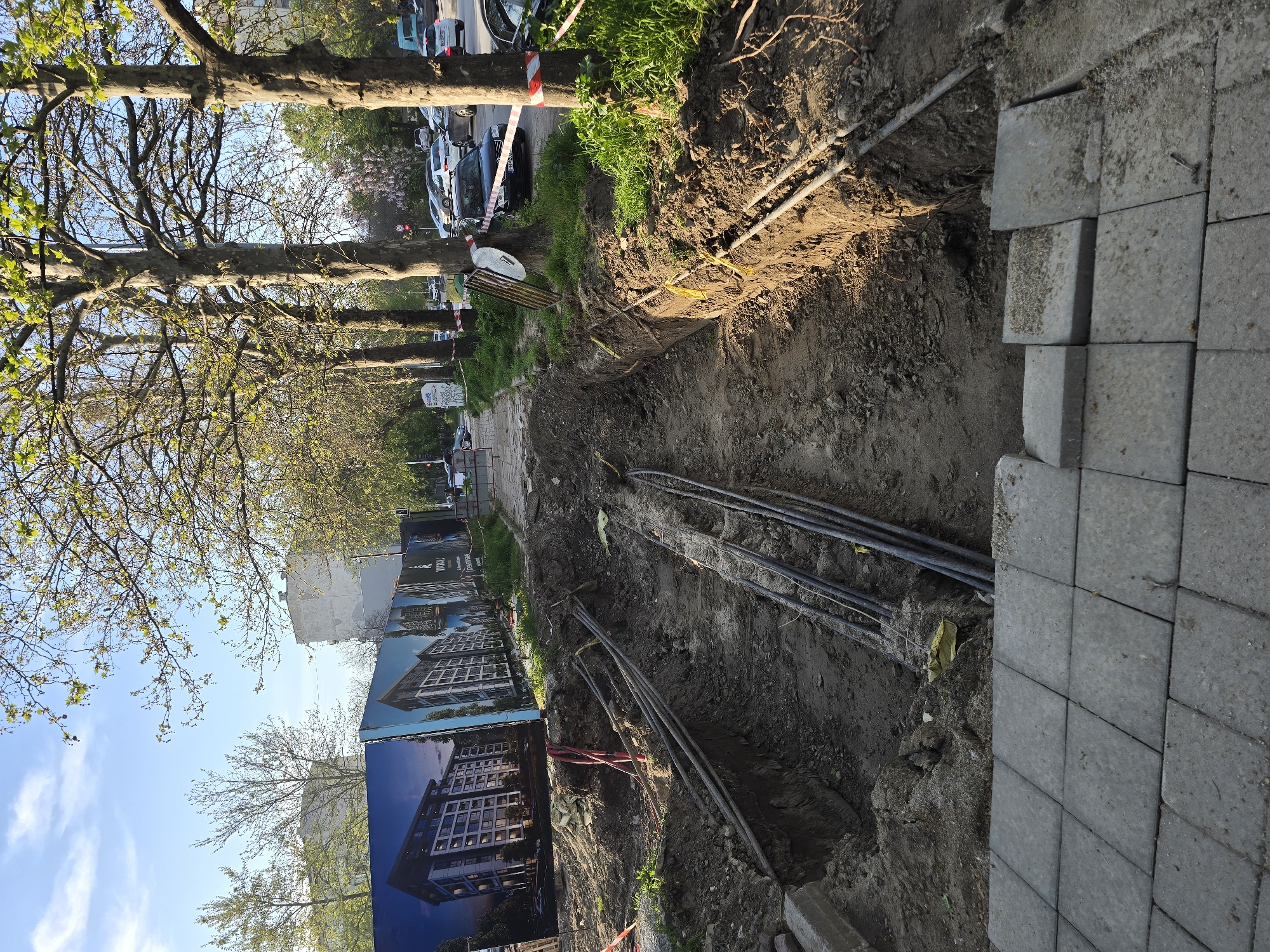 The image depicts a construction site along a sidewalk in an urban environment. A trench has been excavated parallel to the sidewalk, revealing underground cables. A stack of concrete pavers sits adjacent to the trench, likely for sidewalk repairs. A promotional billboard displaying an architectural rendering of a building is visible along the sidewalk. Trees line the street, and parked cars are visible in the background. Red and white striped tape indicates a safety barrier. No people are visible in the scene.