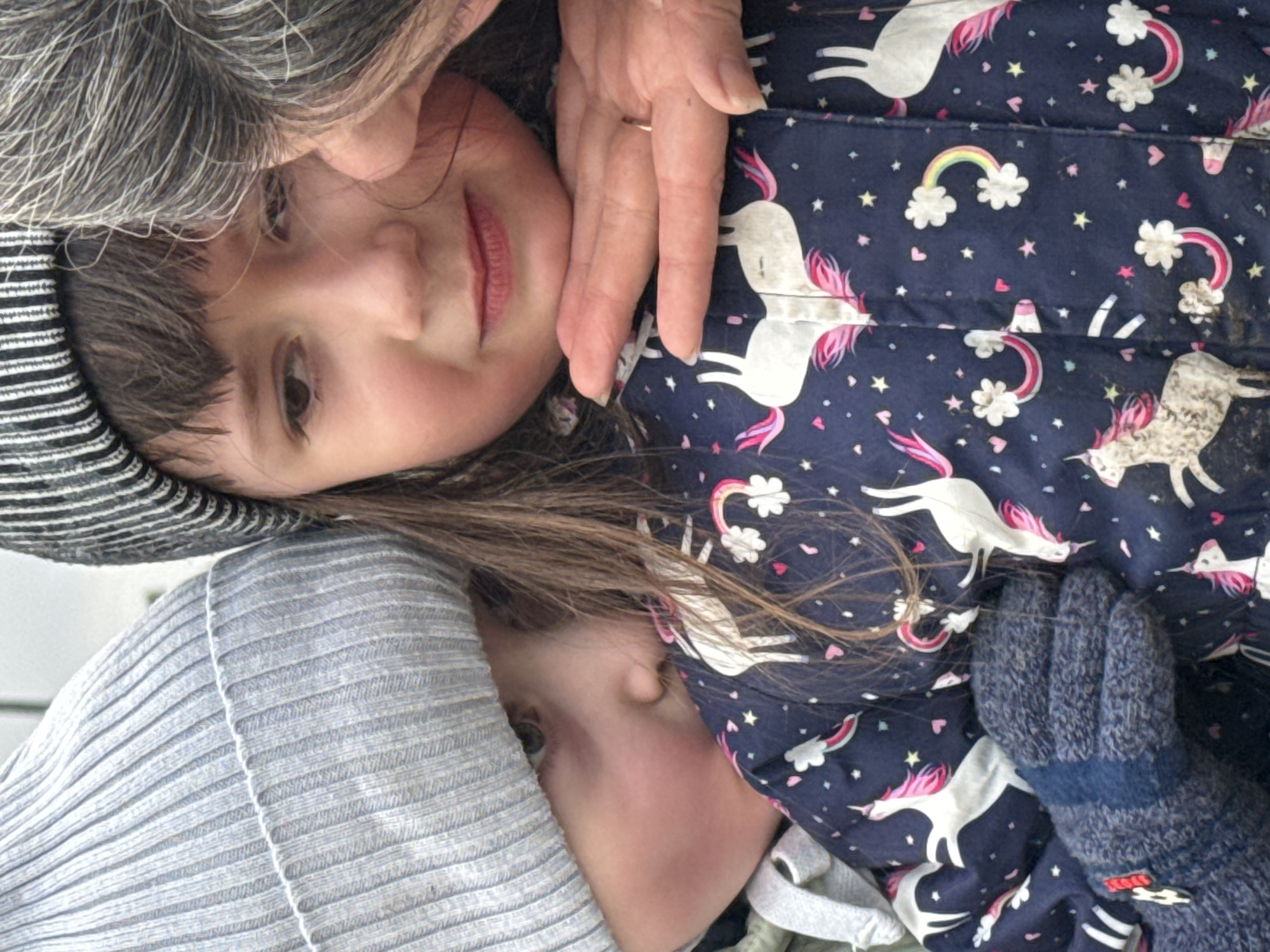 Two young children are lying close together, with an older individual, presumably a parent or grandparent, hovering protectively with a hand near the top child's face. The upper child, wearing a striped hat, is looking up with a faint smile. The lower child is wearing a gray beanie and looking towards the right side of the frame. Both children are bundled in winter clothing. The upper child is wearing a dark blue jacket printed with white unicorns, rainbows, and stars. The lower child is wearing a grey hat. A blue mitten is visible near the right side of the frame. The location and city of Chaikovsky, Russia, are not discernible from the visual information.