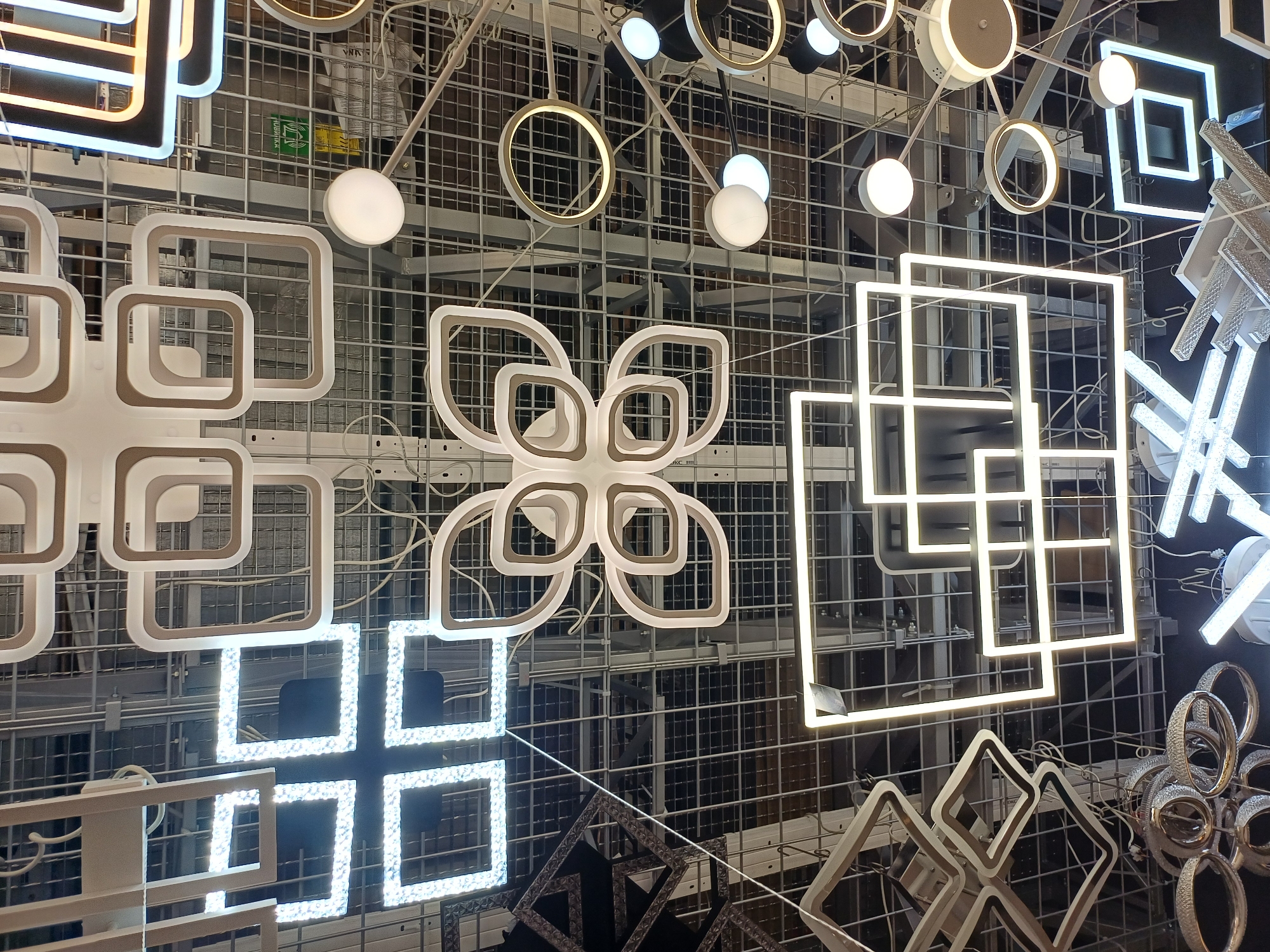 The image shows a ceiling interior with numerous modern light fixtures. The fixtures are predominantly white and geometric in design, with some featuring LED lighting. Several of the lights are square or rectangular, with some interlocking. One fixture resembles a stylized flower or butterfly. The lighting itself varies, with some emitting a bright white light and others a warmer, yellowish hue. The ceiling is constructed with a grid of metal beams and wires, indicative of a commercial space, possibly a showroom or retail store. There is a green and yellow sign with Ukrainian text visible in the upper right quadrant of the image. The overall impression is of a well-lit space showcasing a variety of contemporary lighting options. There are no people or actions depicted in the image. The location of this scene is identified as Boryspil, Ukraine.