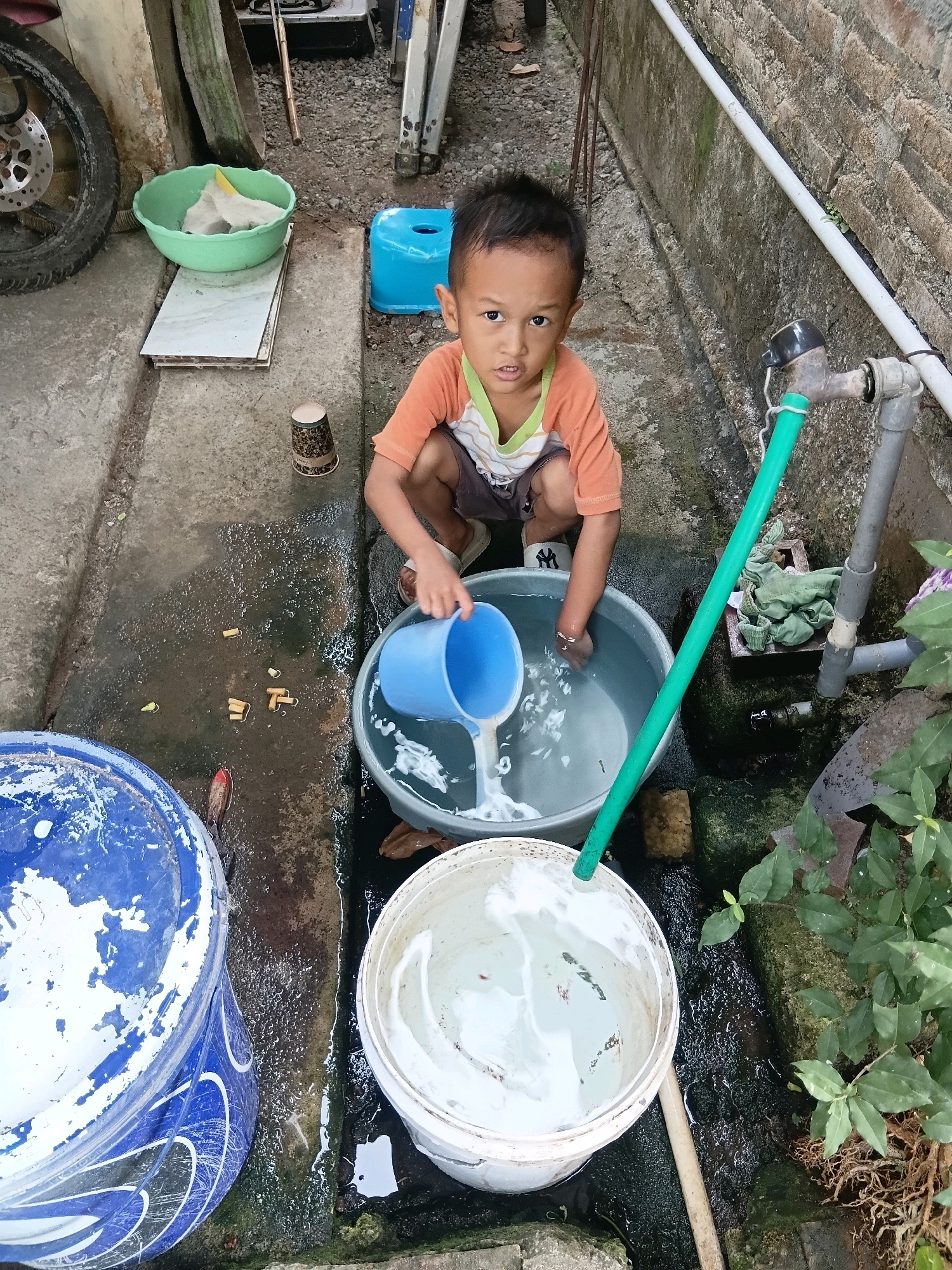 A young boy, likely of Indonesian descent, is seen squatting next to a large gray plastic tub filled with water. He is wearing an orange shirt with a striped collar and dark shorts. In his hands, he holds a blue plastic pitcher, from which he is pouring water into the tub. The scene appears to be an outdoor area, possibly a backyard or alleyway in Cikal, Indonesia, characterized by a concrete ground, a brick wall, and some vegetation. Other objects visible include a blue plastic stool, a green bowl with some items inside, a blue and white painted bucket, a white bucket partially filled with water, a spigot with a green hose, and what appears to be miscellaneous debris on the ground. The overall environment suggests a functional, possibly utilitarian, outdoor space.