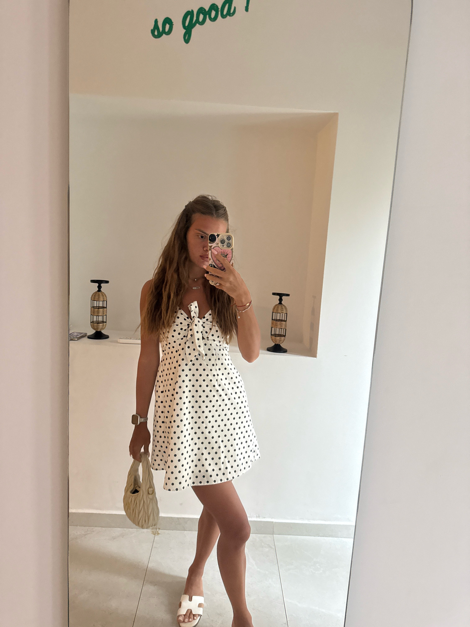 A young woman with long, wavy brown hair is captured in a mirror selfie, standing in a room. She is wearing a white dress with black polka dots, a white handbag, and white sandals. The dress has a bow detail at the bust. The background shows a white wall with the words "so good" written in green cursive script at the top. There are two decorative cylindrical objects on shelves on either side of the mirror. The floor appears to be tiled. The overall ambiance suggests a casual setting, possibly a dressing room or a hotel room in Stavros, Greece.