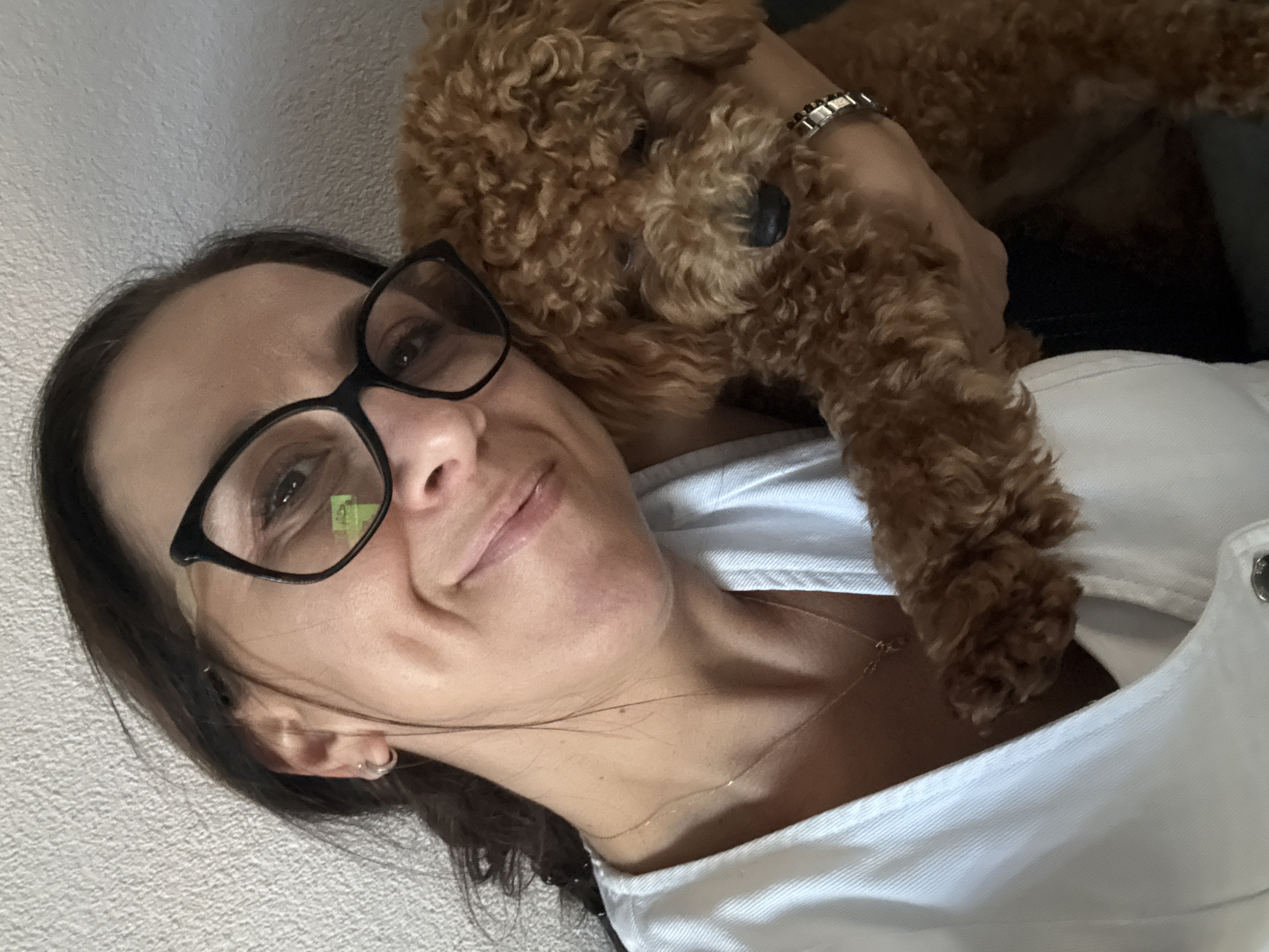 In Marum, Netherlands, a woman with dark hair and glasses holds a small, brown poodle. She is wearing a white shirt and a necklace. The poodle is nestled in her arms, its head resting on her shoulder. The background appears to be a textured wall.