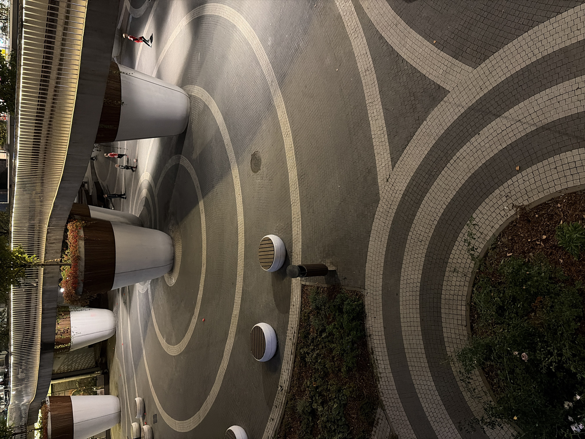 The image captures an outdoor urban plaza at night, featuring a modern architectural design. A raised pedestrian walkway, illuminated by warm LED lights along its railing, spans the upper portion of the frame. Beneath the walkway are large, conical planters with dark wooden accents and white bases, each supporting a small tree. The plaza floor is paved with intricate patterns of dark and light cobblestones, forming concentric circles and flowing curves.
Several people, including children, are visible in the plaza. Some children appear to be playing, with one kicking a red ball. A child in a red outfit is running towards the right side of the frame. The plaza is sparsely furnished with round, white benches with wooden slatted seats. A dark, cylindrical trash receptacle stands near a patch of greenery. The overall atmosphere is one of contemporary urban design with elements of nature integrated into the space, illuminated by artificial lighting. The scene is set in a public urban area, likely a park or a pedestrian zone. There is no information in the image to suggest the location is Moscow, Russia.