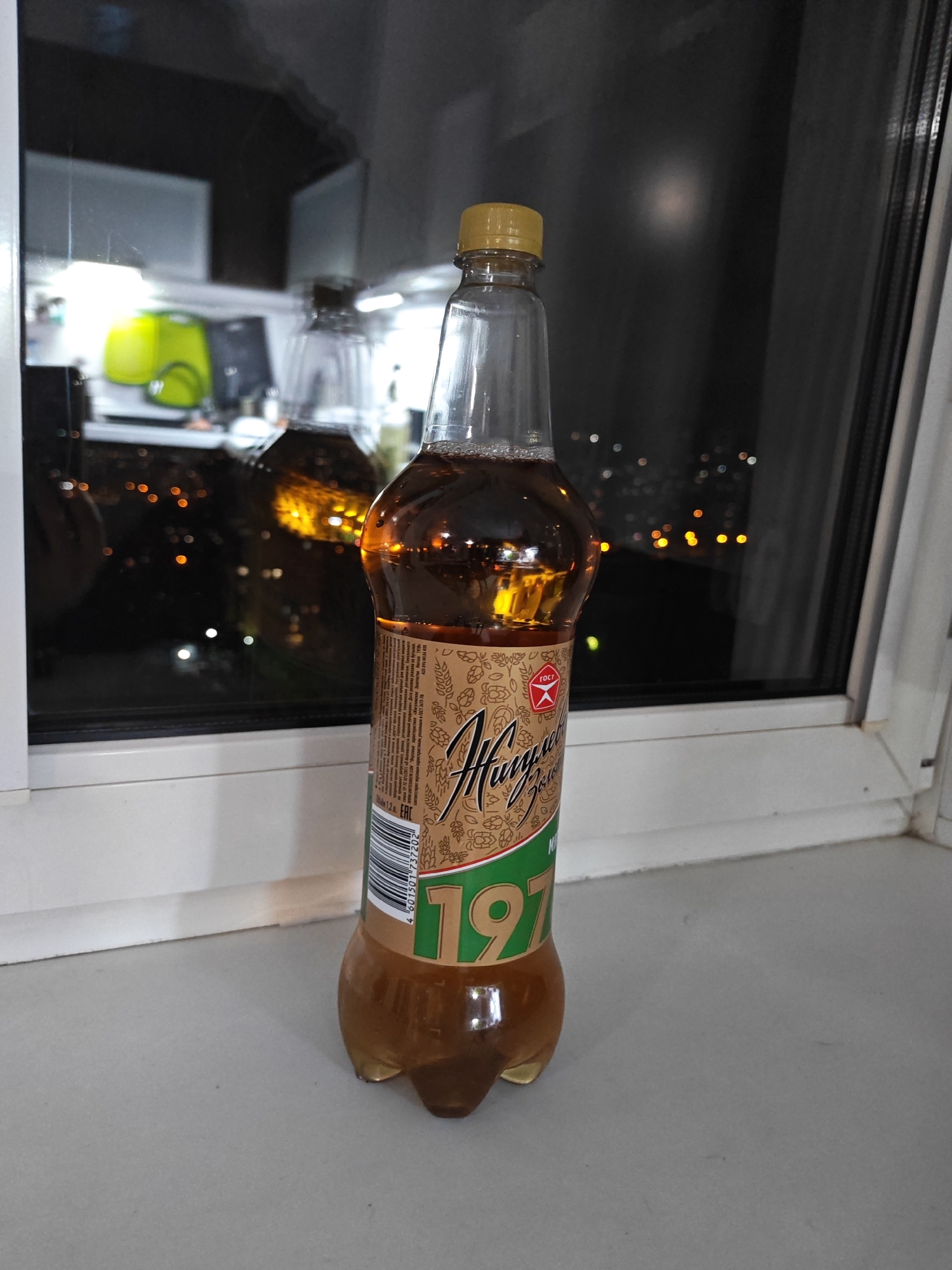 A plastic bottle of "Chernogolovka 197" kvass stands on a windowsill in front of a window at night. The window reflects a blurred interior and shows out-of-focus city lights in the background, suggesting a view from a building in Vladivostok, Russia. The bottle is half-full of amber-colored liquid and has a yellow cap. The label features ornate gold lettering and a green section with the number "197". The windowsill is white, and the surface in front of it is a light gray.
