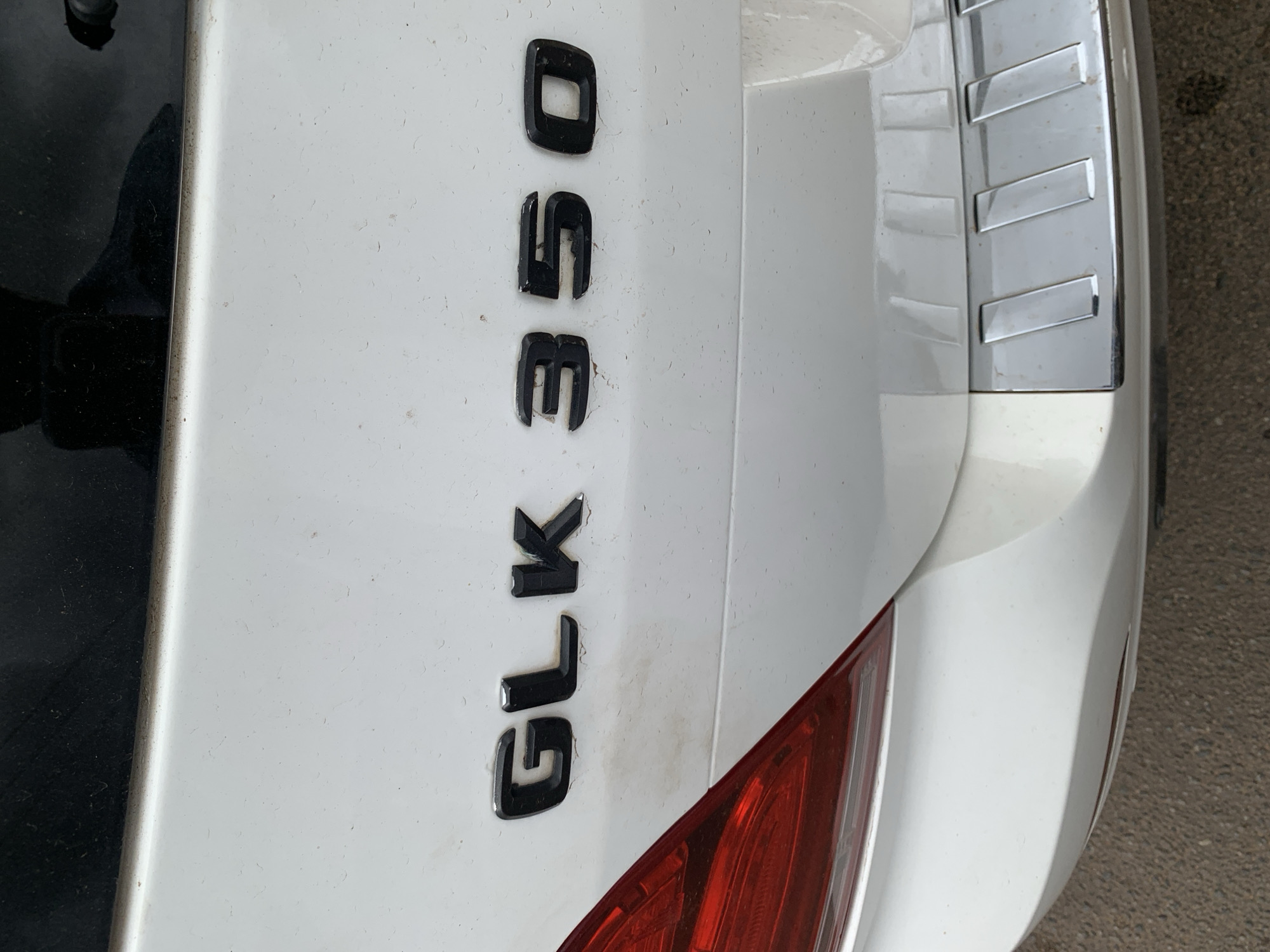 The media file displays a close-up, vertical shot of the rear of a white Mercedes-Benz GLK 350. The vehicle's emblem, "GLK 350," is prominently featured in black, raised lettering on the white car body. To the right of the emblem, a chrome accent strip with horizontal rectangular cutouts is visible, adding a decorative element to the car's design. A portion of a red taillight is also captured in the lower right quadrant of the image.
The car appears to be parked outdoors, with a textured asphalt surface visible in the lower right and right edges of the frame, suggesting a road or parking area. The lighting indicates it is daytime, and the car's surface shows some dust and a few minor scuff marks, typical of a vehicle in regular use. The overall scene is focused on the vehicle's branding and rear design. There are no people or specific events depicted. The location context is identified as Jos, Nigeria.