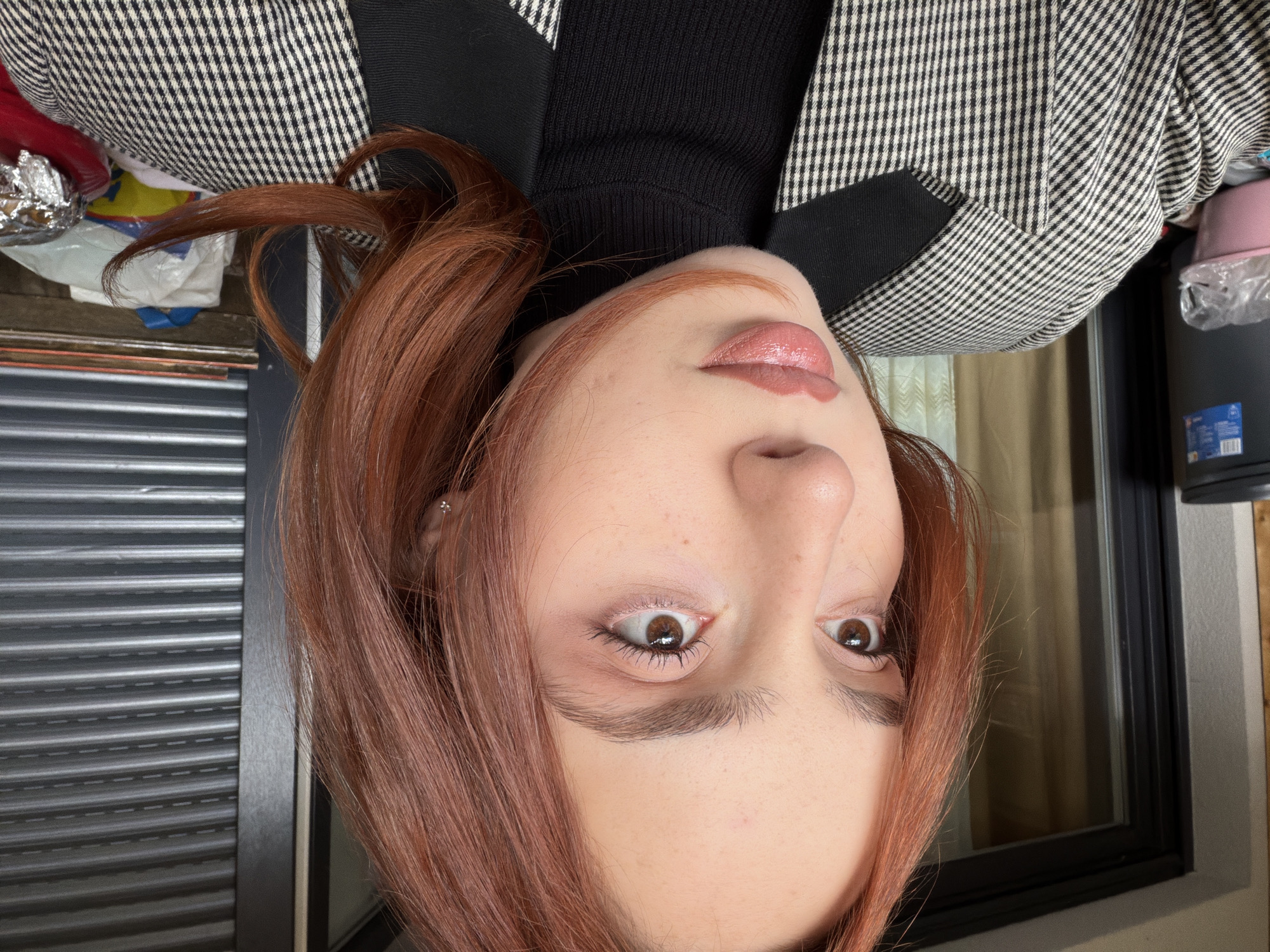 A close-up, tilted image shows a person with auburn hair wearing a black turtleneck and a black and white houndstooth blazer. The person's face is prominent, with a focus on their eyes and lips. The background includes a gray roller shutter, a window, and assorted items like plastic bags and containers. The location and city of Strasbourg, France cannot be determined from the image.