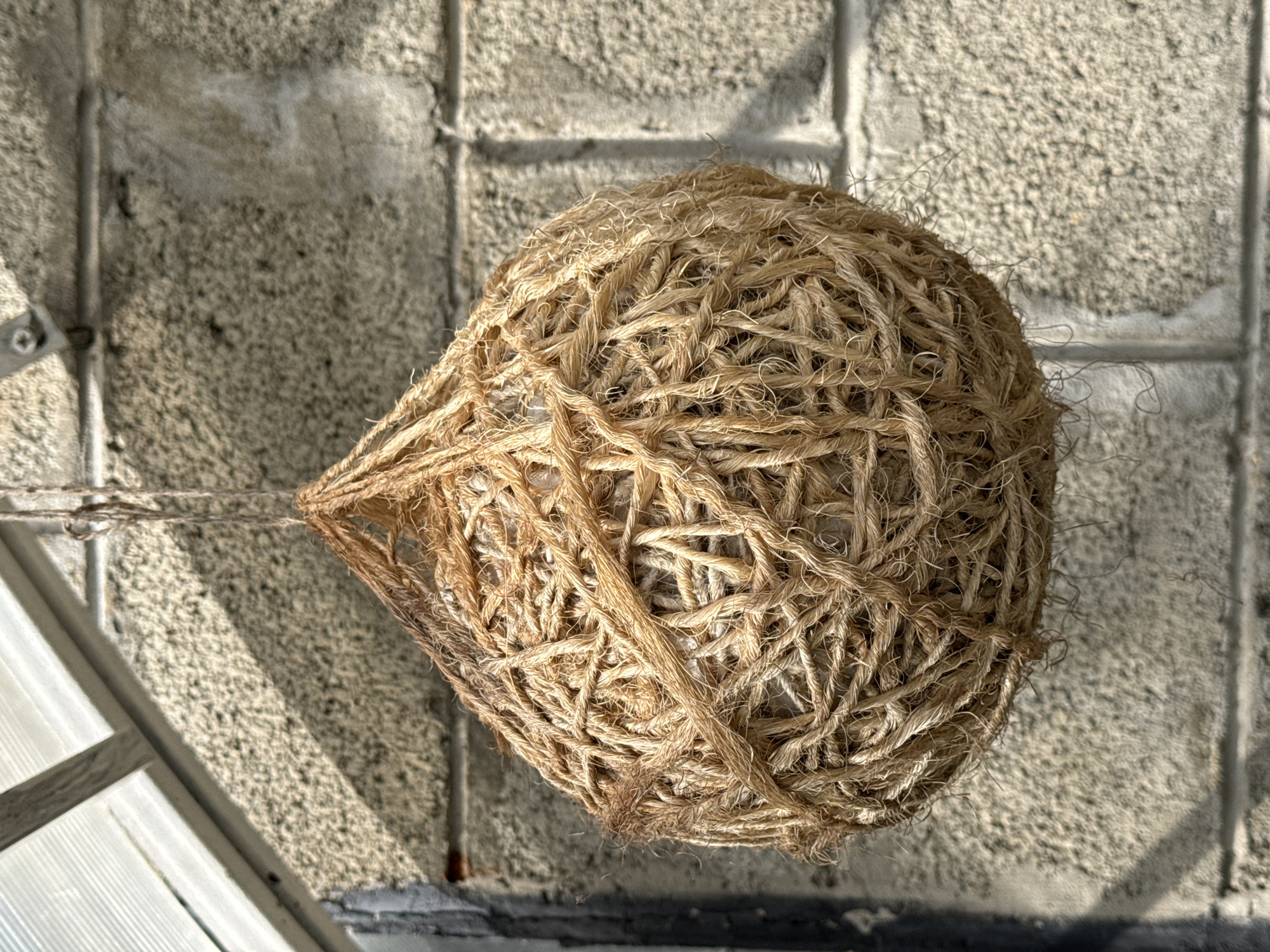 A close-up, eye-level, outdoor shot shows a hanging, woven ball made of rough, brown twine suspended against a light-colored, rough-textured brick wall. The twine is woven in a dense, crisscrossing pattern, forming a spherical shape with an opening at the top. Some lighter-colored material, possibly stuffing or another type of filler, is visible within the twine netting. Sunlight casts shadows across the brick wall, indicating it is daytime. In the upper left corner, a portion of a clear, ribbed roof structure is visible, supported by a metal bracket attached to the wall. The scene appears to be in a garden or greenhouse setting, possibly in a rural or suburban area.