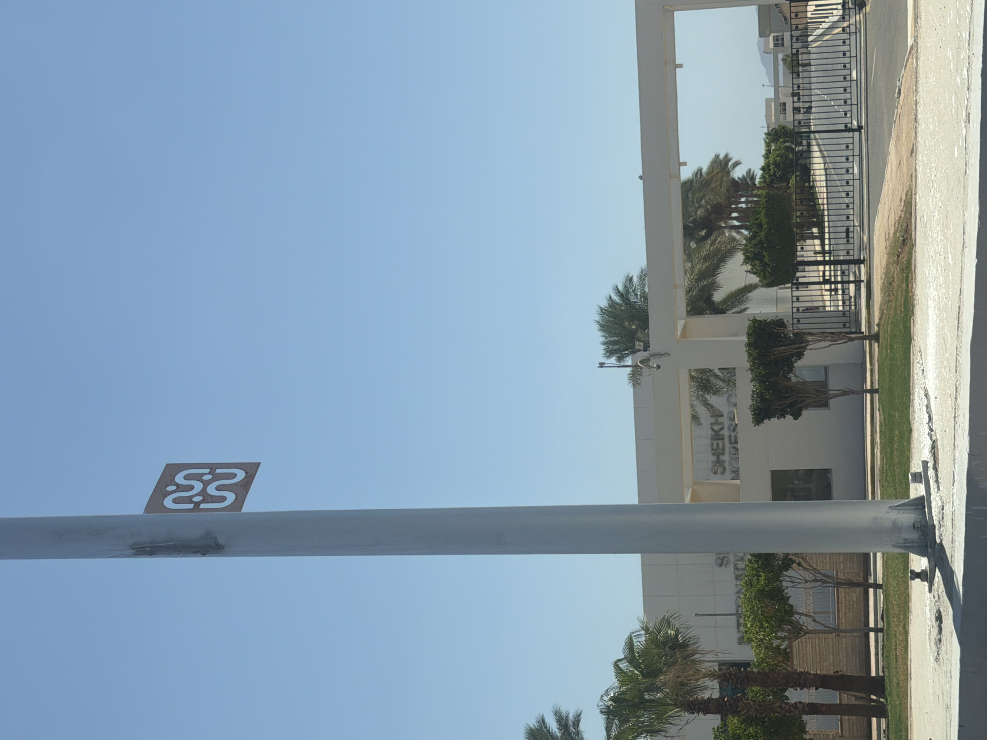 A tall, white pole stands prominently in the foreground, slightly off-center to the left, against a clear blue sky. Attached to the pole, at a moderate height, is a square, brown sign with a white, stylized logo depicting two interlocked "S" shapes. The pole is anchored to a concrete base. Behind the pole, a modern, white, multi-story building with large windows and architectural overhangs is visible. The building has text on its facade, part of which reads "SHEIKH". To the left of the building, palm trees and other greenery are present, suggesting a warm climate. In the middle ground, an entrance gate with black metal bars is closed, leading to a paved road that extends into the distance. Beyond the gate, more trees and buildings are visible, and in the far background, mountains can be seen under the bright sky. The overall scene appears to be an entrance to a facility or complex in Sharm El Sheikh, Egypt.