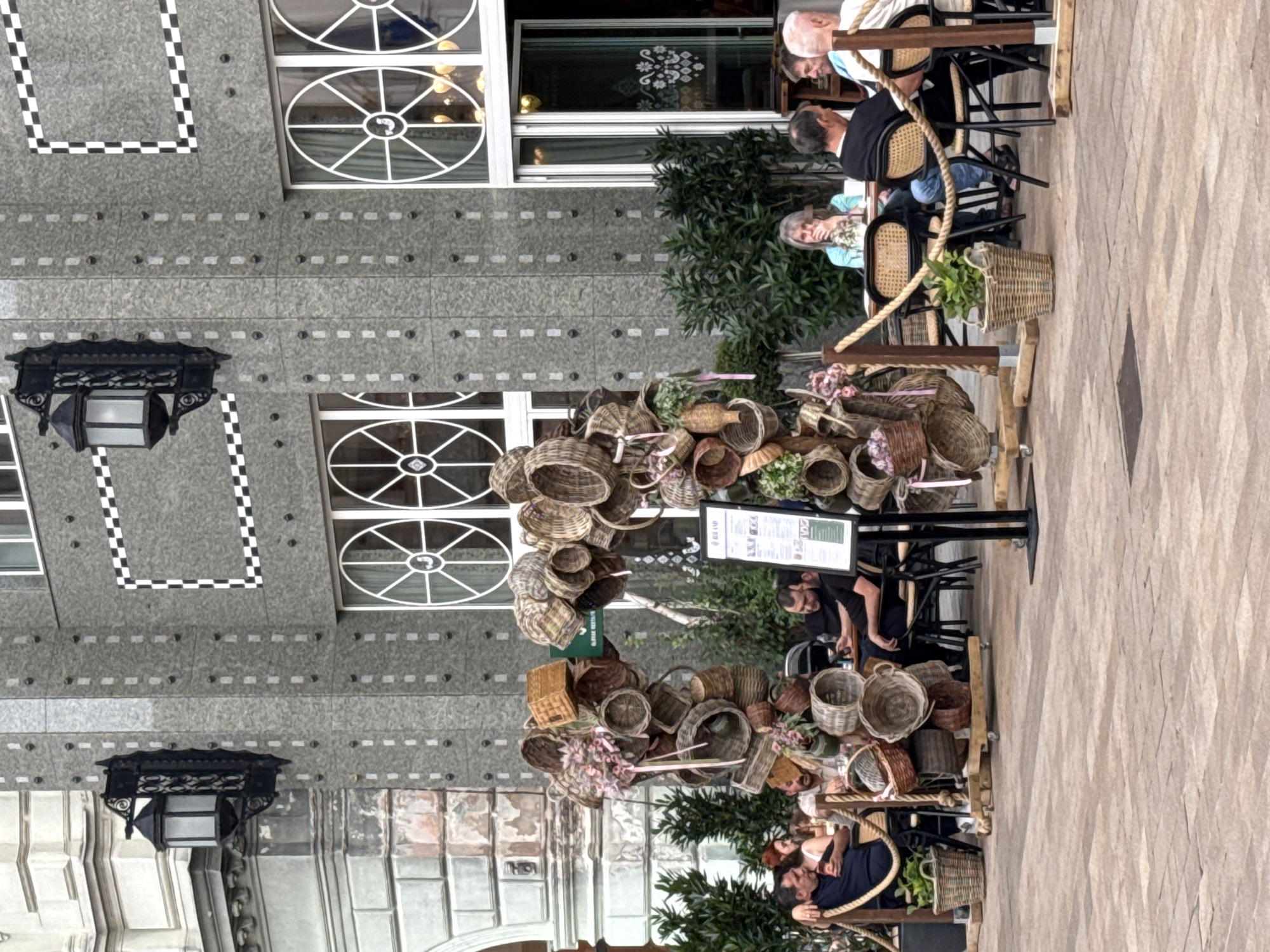 The image shows an outdoor seating area of a cafe or restaurant with several people seated at tables. A large display of wicker baskets is arranged in the foreground. The architecture of the building appears to be ornate, with decorative window frames and textured walls.
In the background, people are seated at tables, some engaged in conversation, while others appear to be relaxing. The setting seems to be a pleasant day, with natural light illuminating the scene. The overall mood is one of casual dining and relaxation.
There are no specific time-of-day cues, but the lighting suggests it is daytime. The weather appears to be fair. The architecture and cobblestone-like pavement hint at a European location, possibly in a historic district. The prominent display of baskets could suggest a market or a shop with a rustic theme.
A sign with some text is visible near the basket display, but the text is not legible. Some people are wearing casual attire, while others are dressed more formally, suggesting a mix of patrons. The overall impression is a lively yet relaxed outdoor atmosphere.