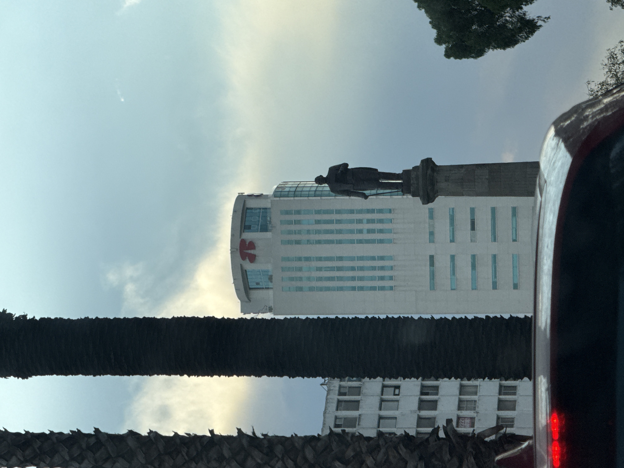 The image shows a statue atop a monument in Puebla, Mexico, framed by a tall building with a red logo, a palm tree, and a portion of a red vehicle. The sky is visible in the background with some clouds. The monument is grey and the statue depicts a man. The building is white with rows of blue windows. A second building is visible behind the palm tree. The image is slightly tilted.