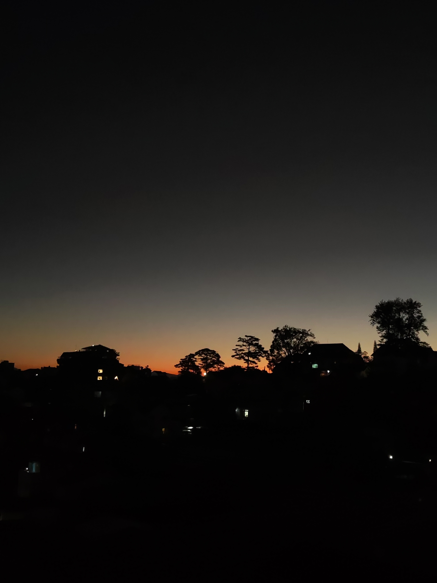 A nighttime cityscape of Da Lat, Vietnam, reveals silhouettes of buildings and trees against a gradient sky transitioning from dark gray to orange on the horizon. The illumination from the sunset is prominent around the tree line. Several buildings have visible window lights, indicating human habitation. The foreground is predominantly dark, obscuring details.