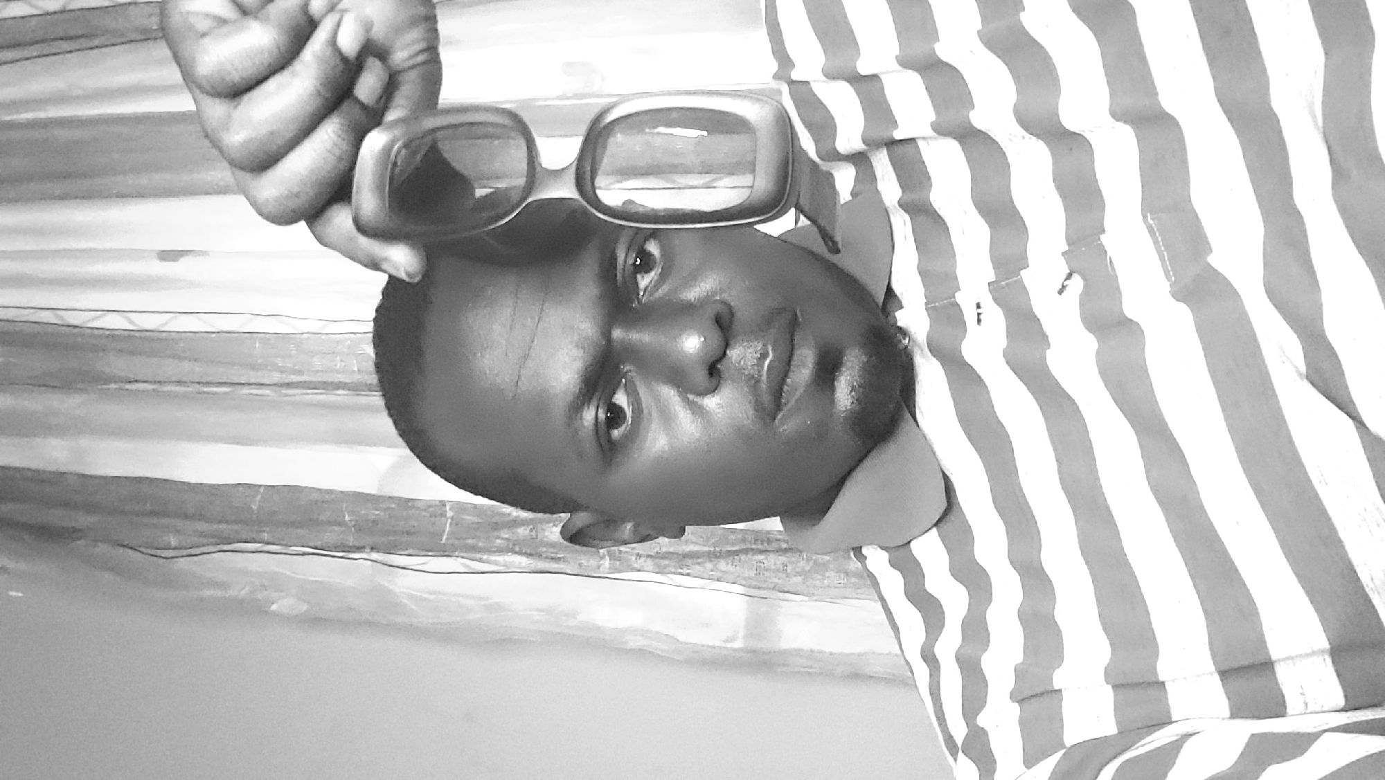The image shows a man, likely in Narayi, Nigeria, based on the prompt. He is holding a pair of sunglasses with his left hand. The man is wearing a striped shirt. The background appears to be a curtain or wall with a similar striped pattern. The image is in black and white.
