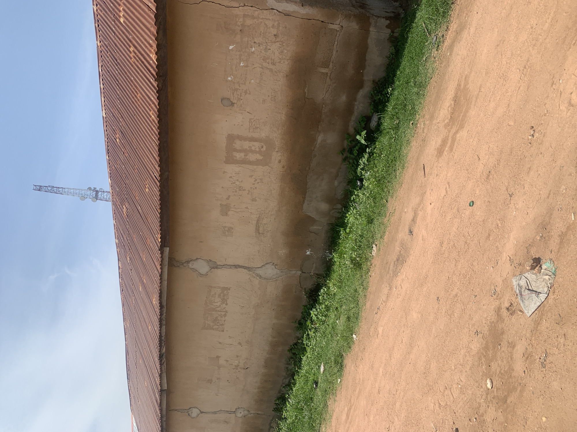 The image shows a sunlit, outdoor scene in Jauro Yinu, Nigeria. A long, weathered wall, made of what appears to be stucco over brick, dominates the mid-ground. The wall is a light brown color and shows signs of age and disrepair, with visible cracks and patches. There are some faint markings or graffiti on the wall, including a square shape and some handwritten text. Above the wall, a rusty, corrugated metal roof is visible, sloping downwards from left to right. In the distance, a tall telecommunications tower with multiple antennas rises against a clear blue sky. The foreground consists of a dirt road, reddish-brown in color, with tire tracks visible. Along the base of the wall, a strip of green grass and weeds grows. A piece of crumpled plastic or fabric lies on the dirt road near the bottom center of the image. The overall impression is of a humble, rural or semi-urban setting.
