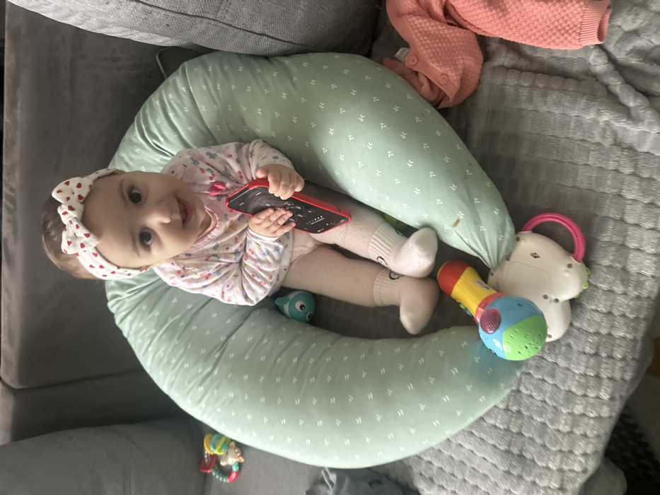 The scene depicts an indoor domestic setting, located in Strasbourg, France. A young infant, with dark hair and eyes, is seated upright within a light-green, U-shaped support pillow patterned with small white geometric shapes. The infant wears a white headband with red hearts and a bow, a long-sleeved light-colored shirt featuring a subtle pattern, light-colored lower garments, and white socks with a dark design near the ankles. The infant holds a red-cased smartphone in both hands, oriented with the screen facing away and appearing dark. The infant is looking directly at the viewer with a slight open-mouthed smile.
The support pillow and infant are positioned on a textured gray blanket or throw, which covers gray upholstered furniture. To the infant's left, a colorful ring-shaped rattle is partially visible. Beneath the infant's legs, resting on the gray blanket against the support pillow, are a multi-colored toy resembling a microphone (red, yellow, blue, green) and a white and pink toy with a handle. A small, light blue and white toy is situated between the infant and the left side of the support pillow. To the infant's right, a folded pink knitted garment is present on the gray blanket. Portions of the gray upholstered furniture are visible in the background on both sides of the frame.