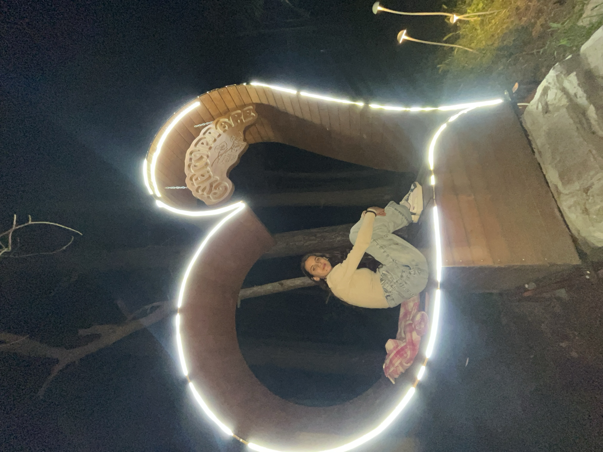 A young woman hangs upside down from a light-lined, wooden structure at night. The structure, which resembles an ampersand and is adorned with carved wooden details at the top, is located in Sungurlare, Bulgaria. The woman is wearing a light-colored shirt, light-wash jeans, and white shoes, and she appears to be holding onto the structure. A pink and white patterned fabric hangs down behind her. In the background, there are illuminated mushroom-shaped lights, trees, and a stone structure. The area is dark, illuminated primarily by the structure's lights.