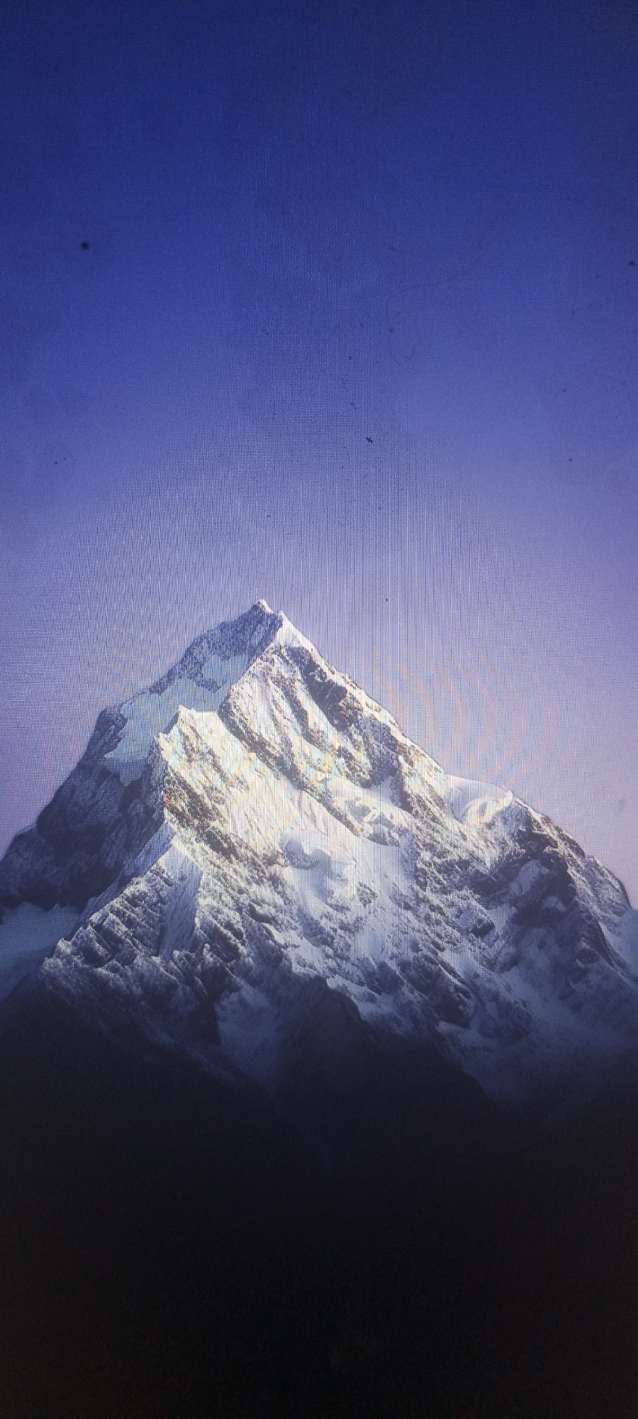 A majestic, snow-capped mountain dominates the frame, its peak reaching towards a clear, deep blue sky. The mountain's slopes are rugged and textured, with patches of snow highlighting its sharp ridges and deep crevasses. Sunlight bathes the upper portion of the mountain, creating a dramatic contrast between the illuminated snow and the shadowed valleys below. The lower part of the mountain fades into darkness, obscuring the base. The image appears to be a photograph or digital rendering. There is no indication of a city location, including Kudymkar, Russia, within the visual content.