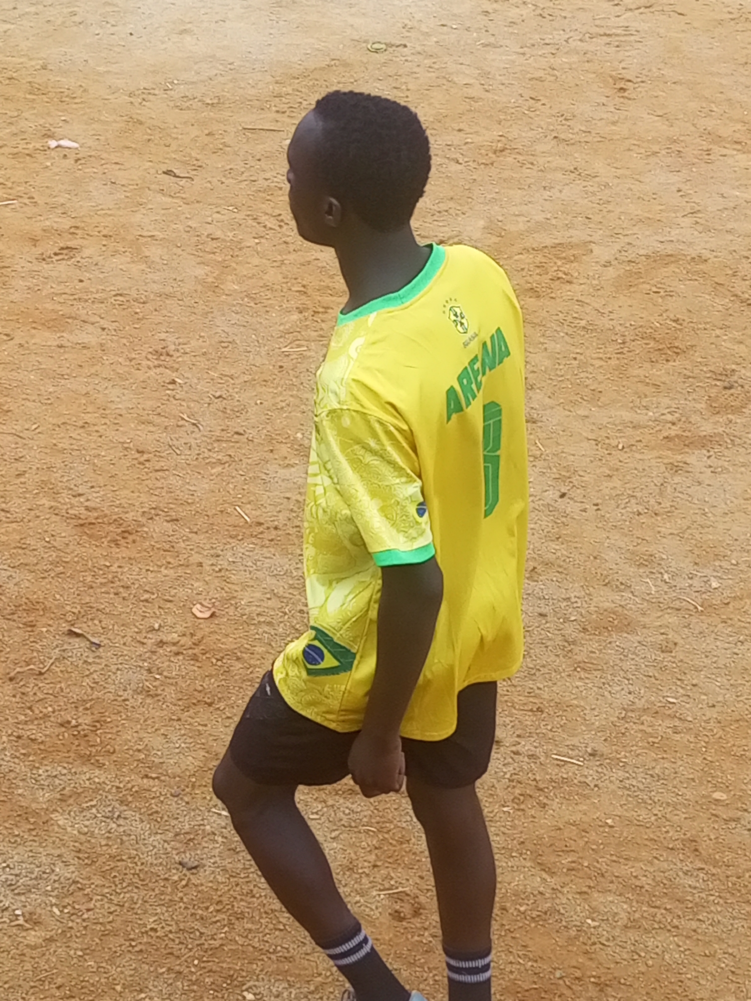 The image shows a young Black male standing on a sandy surface, likely a sports field, in Jos, Nigeria. He is wearing a bright yellow t-shirt with green trim, featuring the word "ARENA" in green lettering on the back and a Brazilian flag emblem on the sleeve. He also wears black shorts and black and white striped socks. The sandy ground is scattered with small pebbles and debris. The lighting suggests it is daytime. The individual appears to be looking away from the camera, towards the right side of the frame.