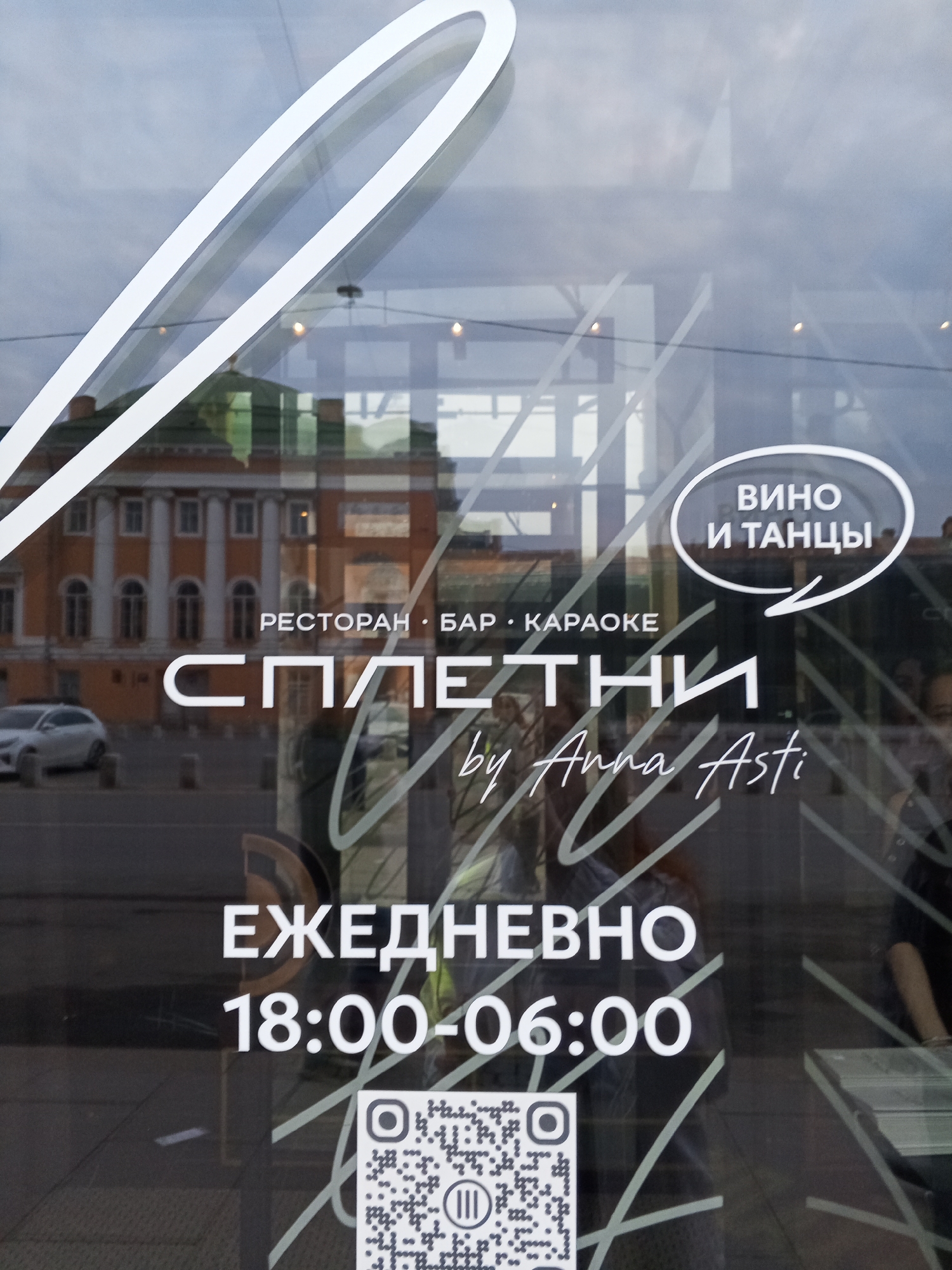 The image shows the exterior of a restaurant, bar, and karaoke venue called "Spletni" by Anna Asti in Saint Petersburg, Russia. The signage on the glass door indicates it is open daily from 18:00 to 06:00. A white speech bubble on the glass states “Вино и танцы" (Wine and Dancing). There is a QR code at the bottom. Reflections in the glass reveal a classical-style building across the street and passersby outside.