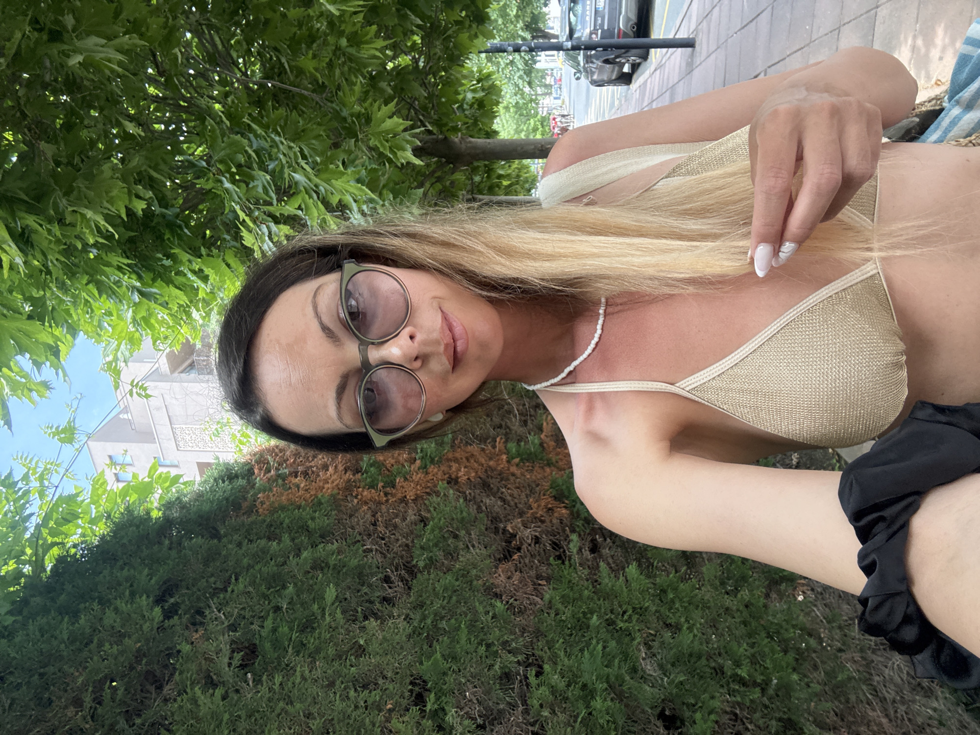 A woman is taking a selfie outdoors, standing next to a hedge and beneath trees. She is wearing a gold bikini top, a white beaded necklace, and sunglasses. Her blonde hair is down, and she is holding a black scrunchie in her left hand. In the background, a building, a sidewalk, a street, cars, and other pedestrians are visible. The scene appears to be in the city of Nesebar, Bulgaria, given the architectural style of the building and the general environment.