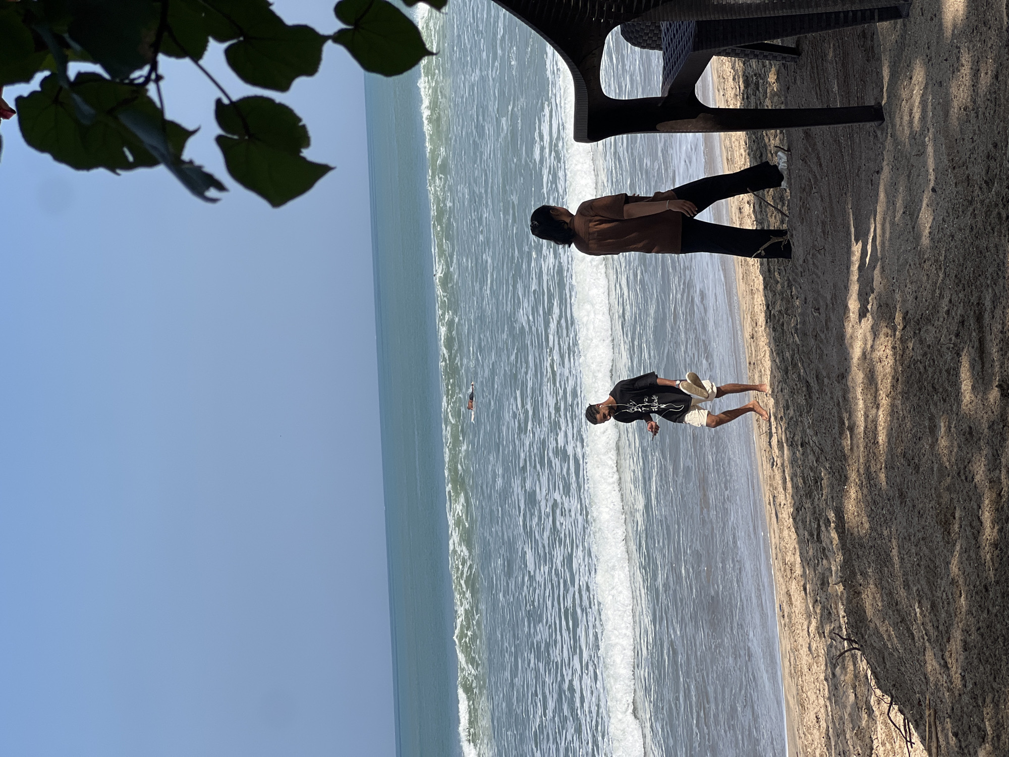 In Kuta, Bali, Indonesia, two individuals are on a sandy beach with the ocean and waves in the background. A man in a black t-shirt and shorts walks along the shoreline, holding a pair of flip-flops. A woman in a brown short-sleeved shirt and dark pants stands near a black plastic chair. In the distance, a person is visible in the water, appearing to be swimming or surfing. The sky is clear and blue, and leaves from a tree hang into the frame from the upper right.