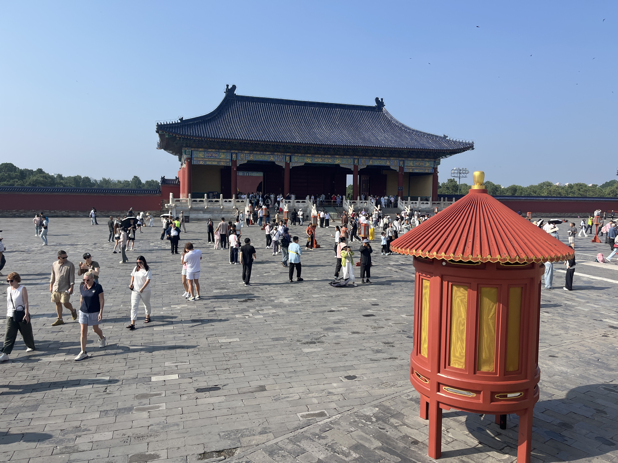 The image shows a wide, paved courtyard in front of a large, traditional Chinese building with a dark blue, tiled roof and red pillars. The building is ornate with decorative yellow and green trim. A bright red, hexagonal lantern-like structure with yellow panes stands in the foreground on the right. Numerous people, dressed in casual summer clothing, are scattered throughout the courtyard, some walking, some standing, and some taking pictures. The sky is clear and blue, with a few birds flying in the distance. The paving stones are large and rectangular, laid in a herringbone pattern. The scene appears to be a tourist destination on a sunny day.