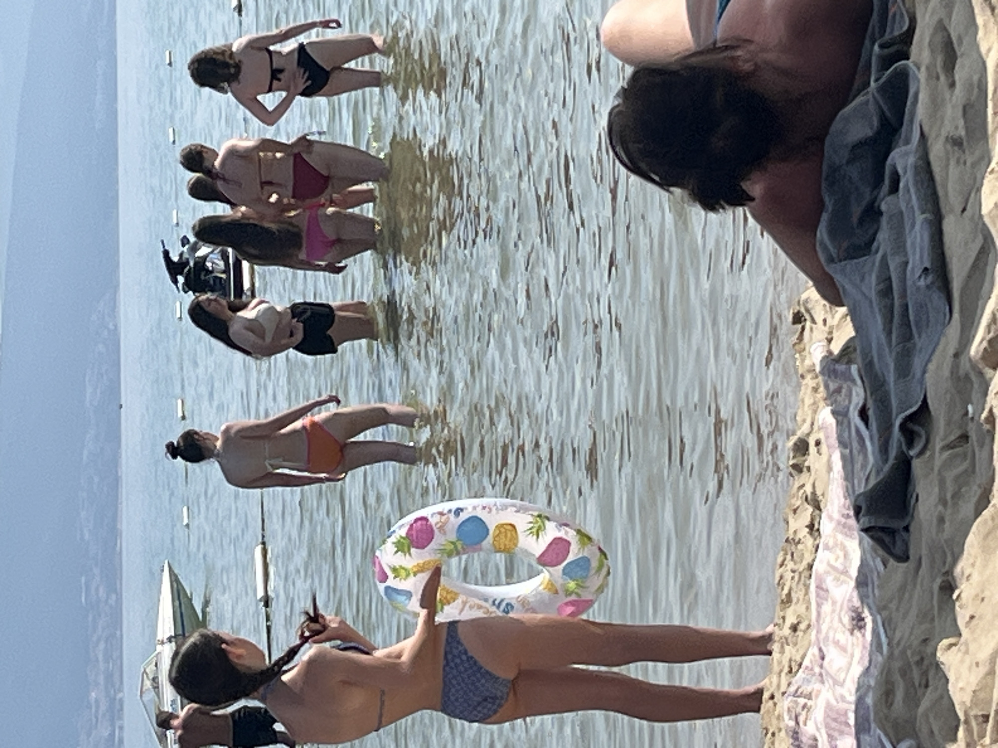 The image depicts a crowded beach scene with people in the water and on the sand. In the foreground, a young woman wearing a patterned bikini bottoms and a dark bikini top is holding a colorful inner tube with a pineapple design. To her right, another young woman in an orange bikini stands in the water. Further back, a group of women in swimwear are wading in the water. A jet ski is visible in the background, along with a boat and a distant shoreline with hills. On the sand in the foreground, towels are laid out, and a person with long dark hair is lying down. The overall impression is a sunny day at a popular beach location.