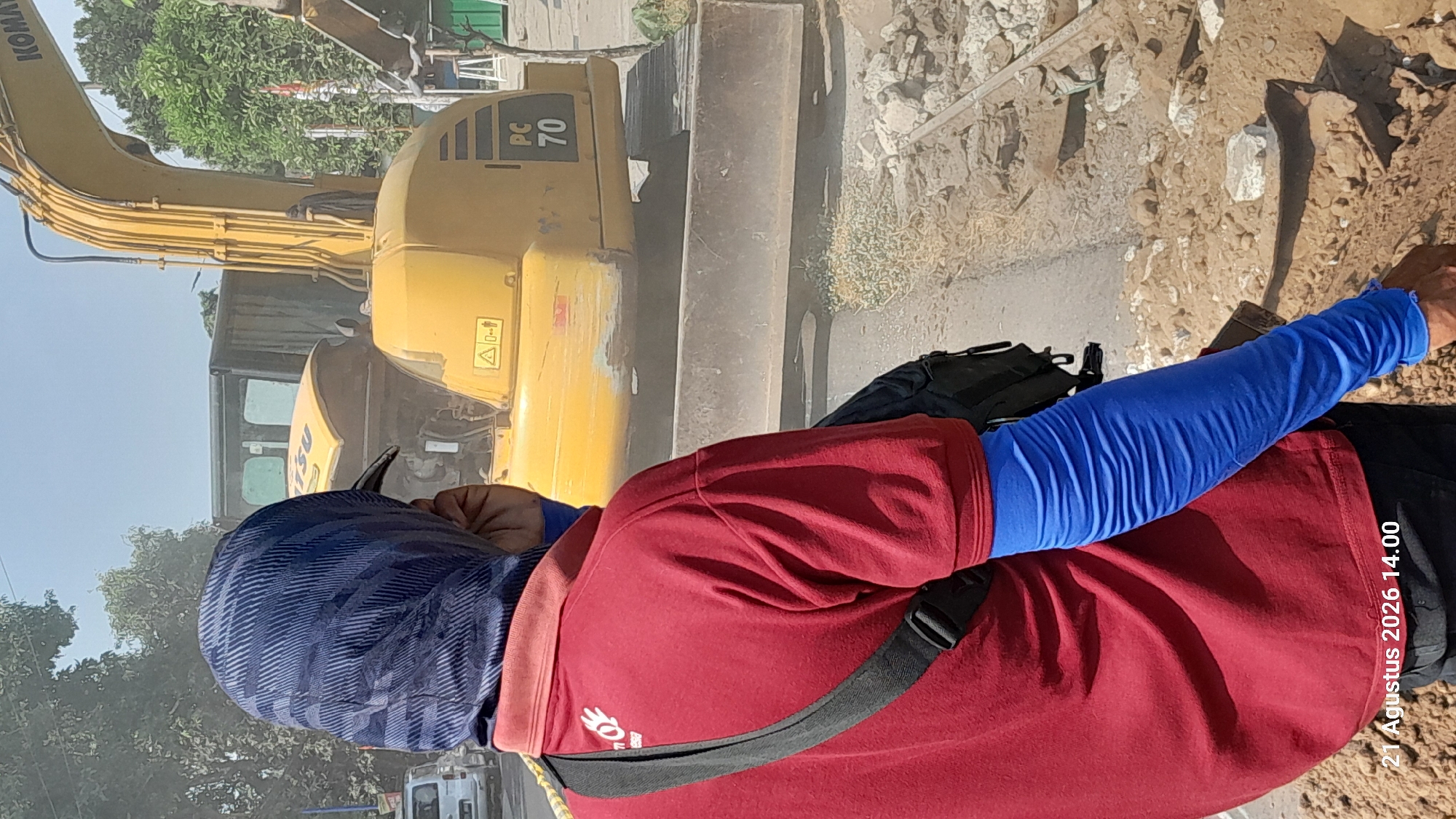 A person wearing a red shirt and a blue headscarf stands facing away from the camera, looking at a yellow excavator on a dirt road. The excavator has the designation "PC 70" on its side. The person has blue arm sleeves pulled up over their arms and is carrying a black bag over their shoulder. The background shows trees and a building. A timestamp "21 Agustus 2026 14.00" is visible in the lower left corner, suggesting the image was taken in Takeran, Indonesia.