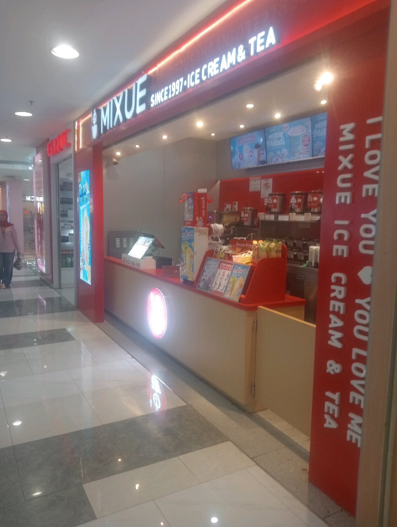 The image shows a MiXUE ice cream and tea shop located in a mall in South Jakarta, Indonesia. The shop has a prominent red and white exterior with a large sign that reads "MiXUE SINCE 1997-ICE CREAM & TEA". Inside the shop, there is a counter with a cash register, a menu displayed on screens, and various ice cream and tea products. A person in a pink hijab and a light pink jacket is walking past the shop on the left side of the image. The floor of the mall is tiled with a combination of white and grey tiles, and there are several ceiling lights visible. The overall scene depicts a typical commercial setting within a shopping mall.