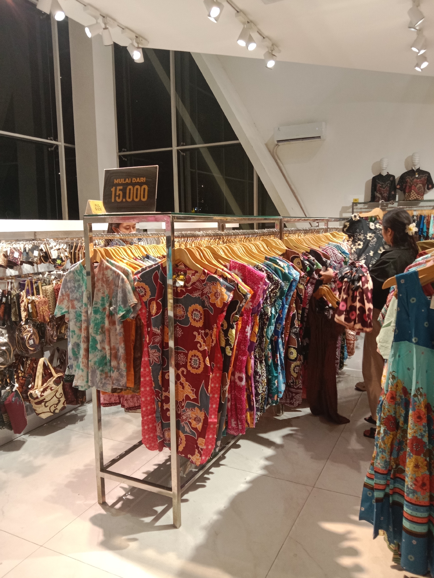 The image shows a retail clothing store interior with racks of colorful batik-patterned clothing. A sign indicating "MULAI DARI 15.000" (Starting from 15,000) is prominently displayed. To the left, there is a rack of smaller items, possibly bags or accessories. In the background, a person is visible wearing a mask and looking at the clothing. Another person, a woman with her hair tied back and a flower accessory, is seen browsing through clothes. Mannequins wearing batik shirts are also visible on a higher rack in the background. The store features modern lighting fixtures and large windows, suggesting a contemporary retail space. The presence of batik clothing strongly suggests the location is in Indonesia, and given the context, it is likely within Semarang, Indonesia, a city known for its batik production.
