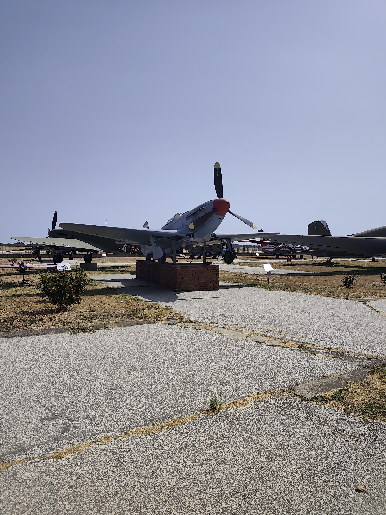 In the city of Krumovo, Bulgaria, an outdoor aircraft museum displays several vintage airplanes under a clear blue sky. In the foreground, a grey and blue propeller airplane, identified as a Lavochkin La-5, is prominently positioned on a brick pedestal. Its propeller is visible, with the nose cone painted red. Behind this aircraft, another plane with olive green wings and a red star insignia on its tail is partially visible. Further in the background, other aircraft are lined up, including what appears to be a dark-colored military jet on the right. The ground is paved with asphalt that shows signs of cracking and is partially covered with dry grass and weeds, indicating an outdoor setting. The overall scene captures a collection of historical aircraft in an open-air environment.