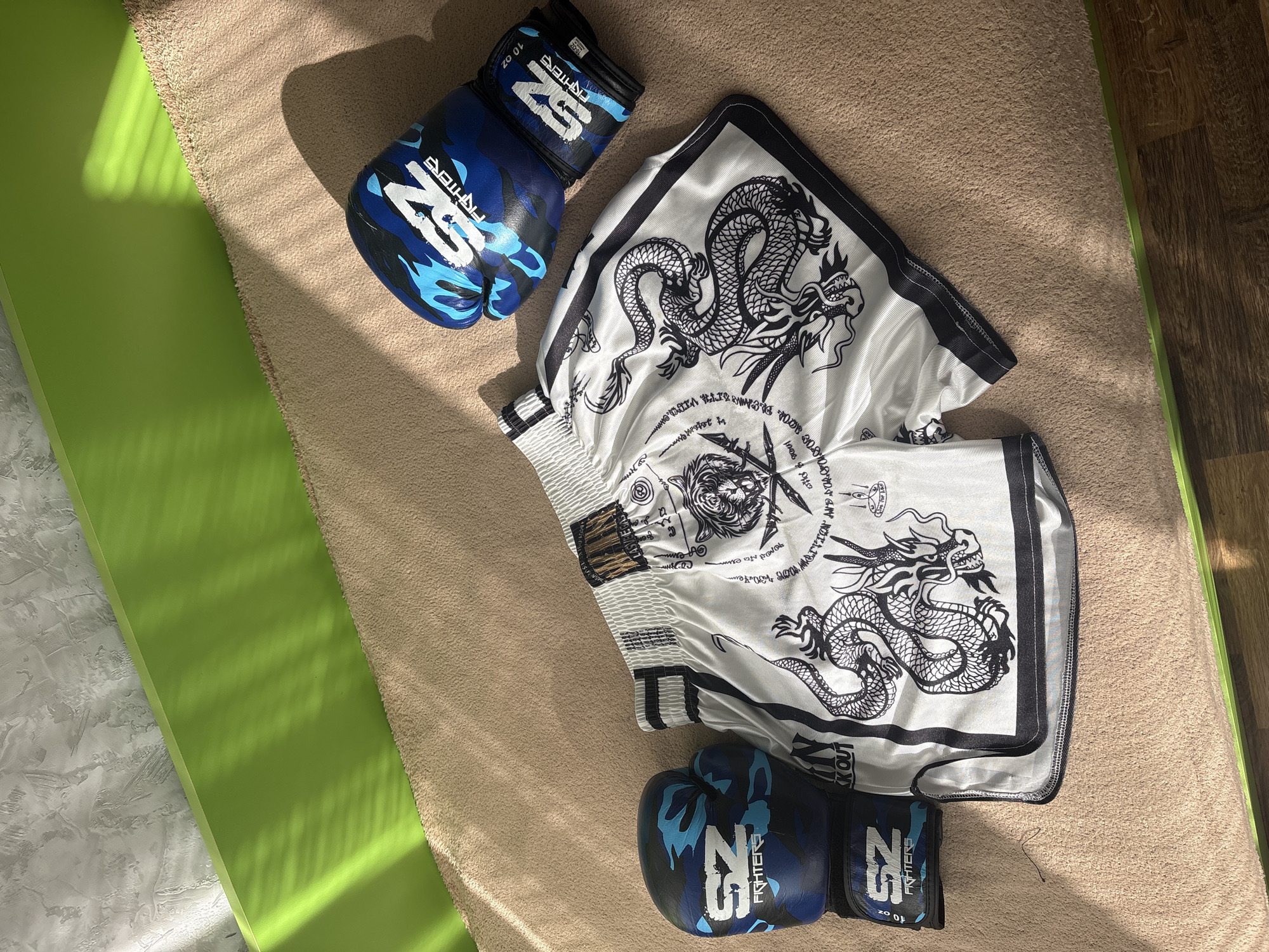 The image shows a pair of blue camouflage boxing gloves and a pair of white Muay Thai shorts with black trim and a dragon design. The gloves are labeled "ZS FIGHTERS" and "10 oz". The shorts also have various inscriptions. All items are lying on a beige towel. A green surface is visible in the corner. The location is not discernible from the image.