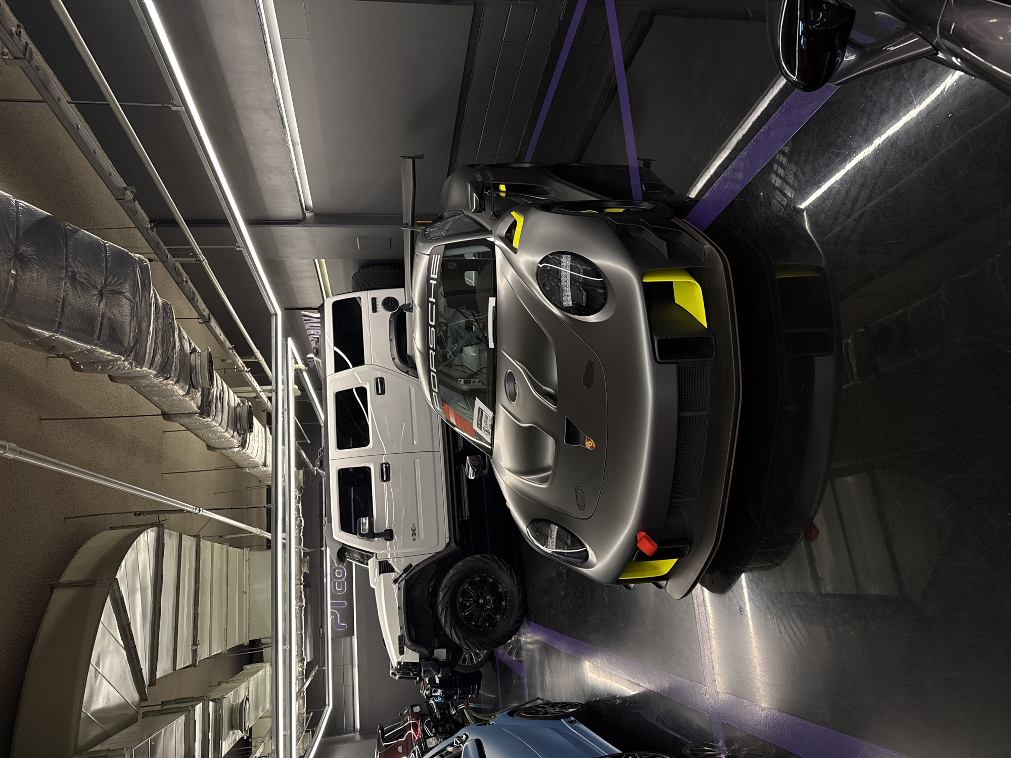 The image depicts a well-lit indoor space, likely a garage or showroom in Seoul, South Korea, given the presence of "PICO" branding on the wall in Korean. A white Jeep-style vehicle and a gray Porsche sports car are parked side-by-side on a dark, reflective floor. Other luxury vehicles are partially visible in the background. Fluorescent lighting fixtures run along the ceiling, alongside industrial ductwork. The scene suggests a high-end automotive display.