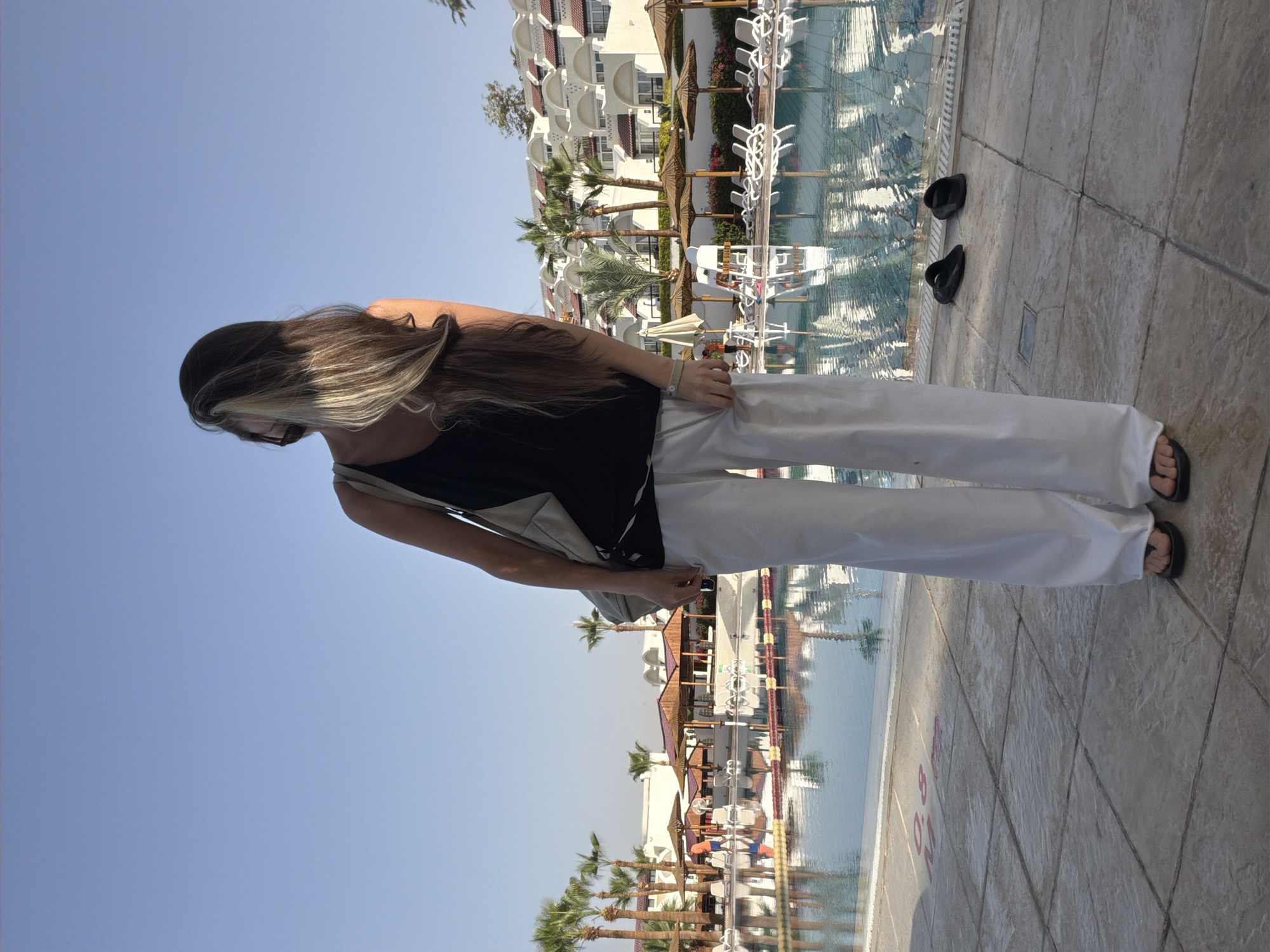 The image shows a woman standing near a swimming pool in what appears to be a resort setting. She is wearing a black top and white pants. Her hair is long and brown with some blonde streaks. She has a purse slung over her shoulder and is wearing flip-flops. Two pairs of black flip-flops lie on the tiled pool deck. The background features a building with white architecture, palm trees, and other resort-style amenities. The water in the pool reflects the sky and buildings. The scene suggests a location in a sunny, vacation-oriented area, consistent with Sharm El Sheikh, Egypt.