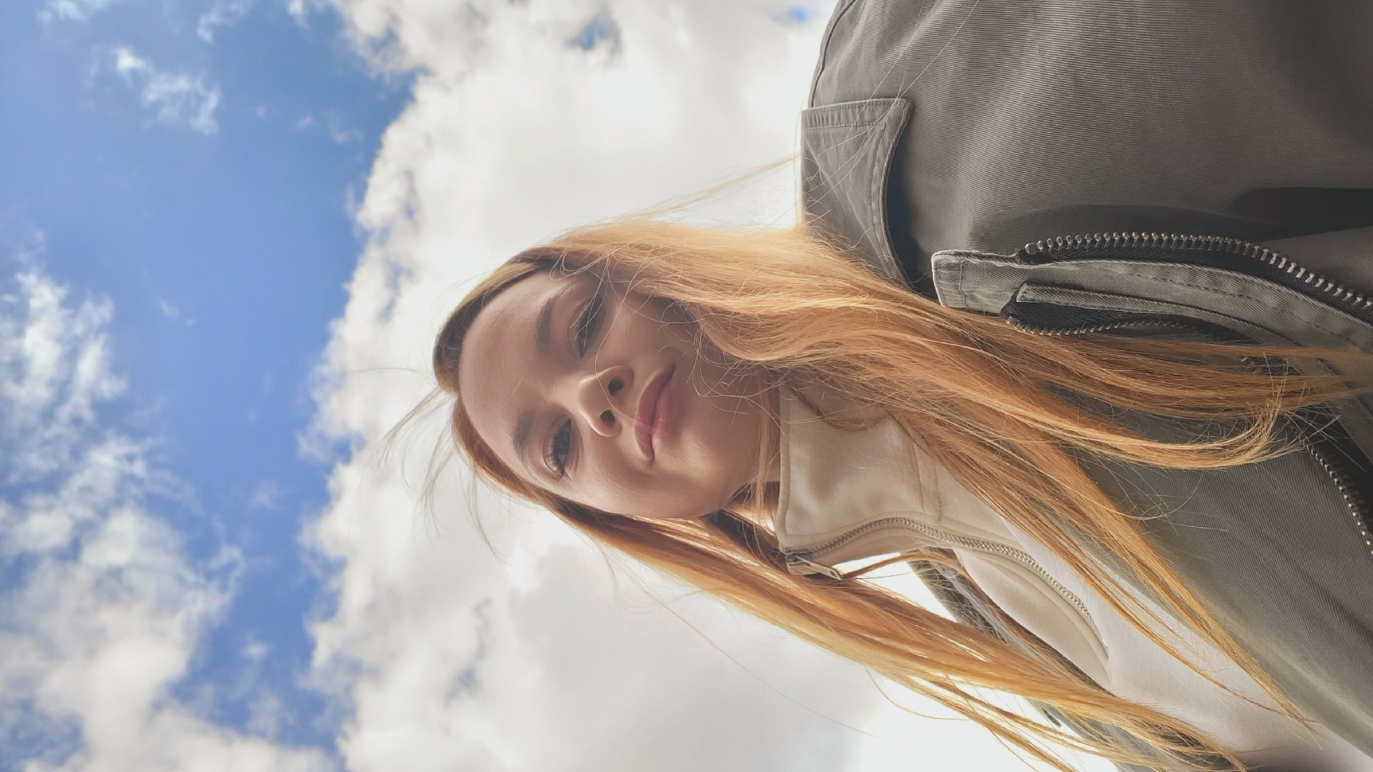 The image, captured in Chaikovsky, Russia, presents a young woman from a worm's-eye perspective, looking down directly at the camera. She has long, wavy, reddish-brown hair that frames her face and flows over her shoulders. Her complexion is fair, and her expression appears neutral or softly contemplative, with her lips slightly parted. She is dressed in a light-colored top, possibly white or off-white, worn under a sturdy olive-green or grey jacket featuring prominent zipper details on the front.
The background is dominated by a bright blue sky dotted with scattered white cumulus clouds, indicating clear, daytime weather. The unusual upward angle suggests that the camera was placed below her, possibly on the ground, as she looked down. No visible text is present in the media file.