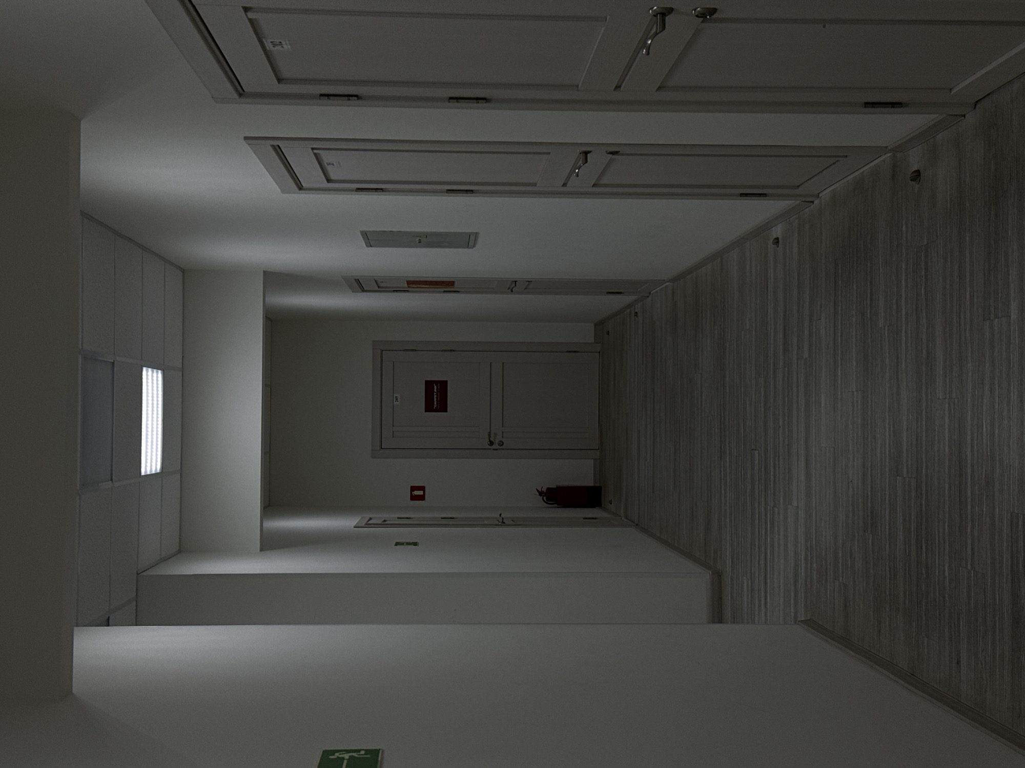 The image depicts a dimly lit, narrow hallway with white walls and a dark wooden floor. Several doors are visible along the right wall. On the left wall, an illuminated green exit sign is present. Further down the hallway on the left, a red fire extinguisher stands beside a door. The hallway appears to be in an institutional or office setting, with fluorescent lighting overhead. No people are present in the image. The overall atmosphere is subdued and somewhat sterile.