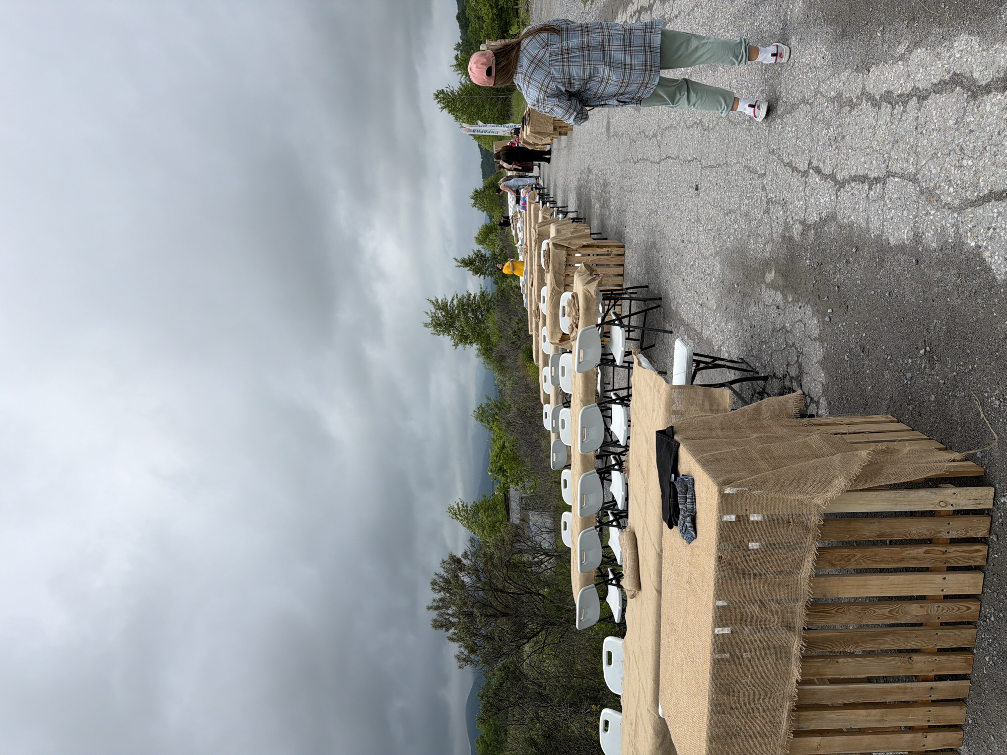 The image depicts an outdoor setting with multiple tables set up, covered in burlap. White chairs are arranged around some of the tables. In the background, there are trees, hills, and a cloudy sky. Several people are visible in the distance, appearing to be interacting around some of the tables. A young woman wearing a plaid shirt, light green pants, and a pink cap is standing in the foreground, facing away from the camera. The scene suggests an event or gathering is about to take place or is in progress. The ground is asphalt, with some cracks and patches of moisture. There is no discernible information to suggest the location is the city of Теплоозёрск, Russia.