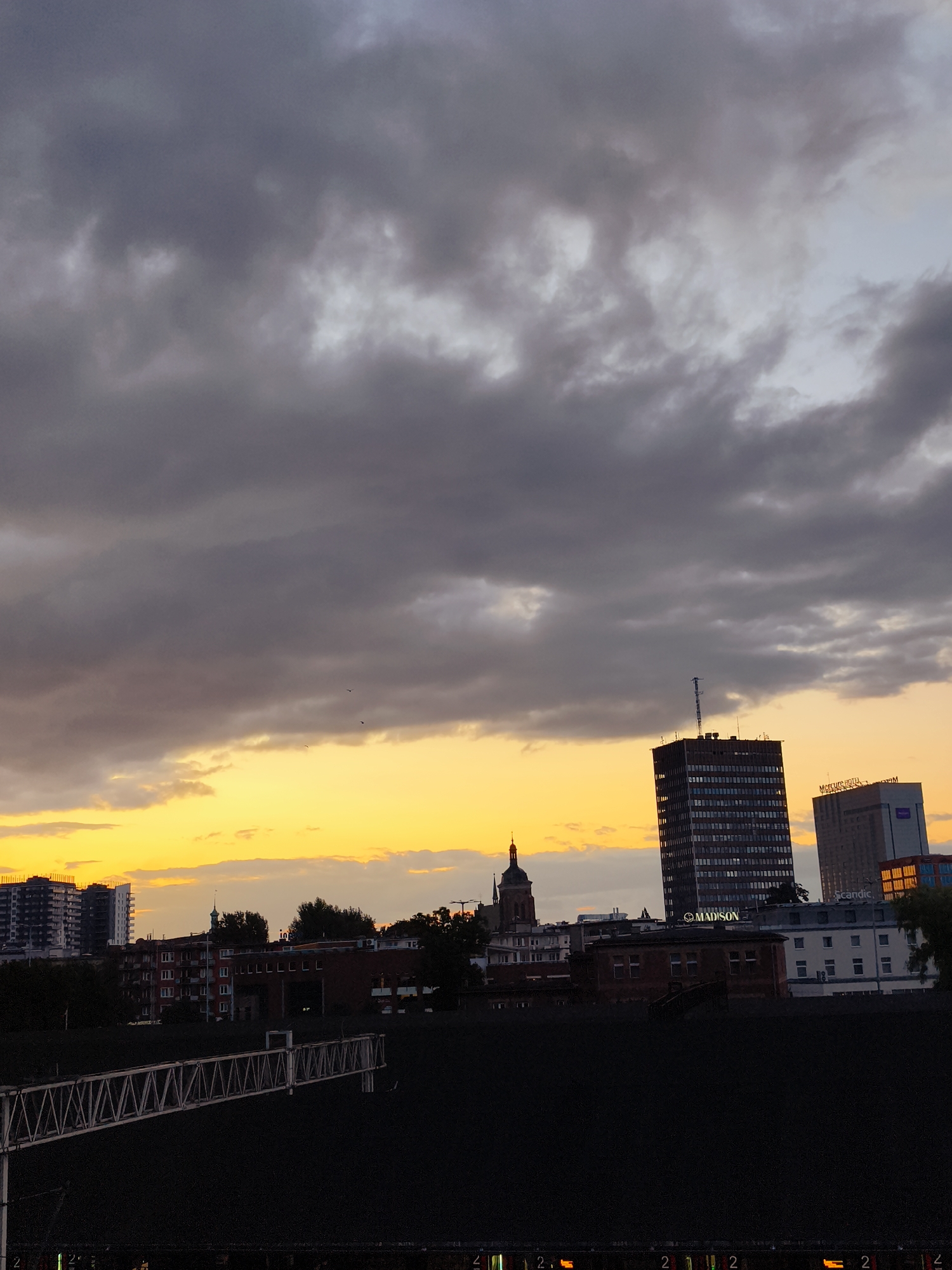The image captures a cityscape of Gdańsk, Poland, at sunset. Dark, dramatic clouds dominate the upper portion of the frame, while a vibrant band of yellow and orange light stretches across the horizon, illuminating the lower sky. Several buildings of varying heights are visible against this backdrop. A tall, dark, rectangular skyscraper with a visible antenna stands prominently in the middle ground. To its right, a lighter-colored building displays signage that reads "Mercure Hotel" and "Scandic." To the left of the skyscraper, a domed structure, possibly a church tower, is partially visible. In the foreground, a dark, textured area, possibly a rooftop or an industrial structure, is present, with the skeletal framework of a large white structure, resembling a bridge or a crane, extending horizontally across it. A few distant birds are faintly visible in the sky. The overall impression is a transition from day to night in an urban environment, marked by atmospheric cloud cover and the warm hues of a setting sun.