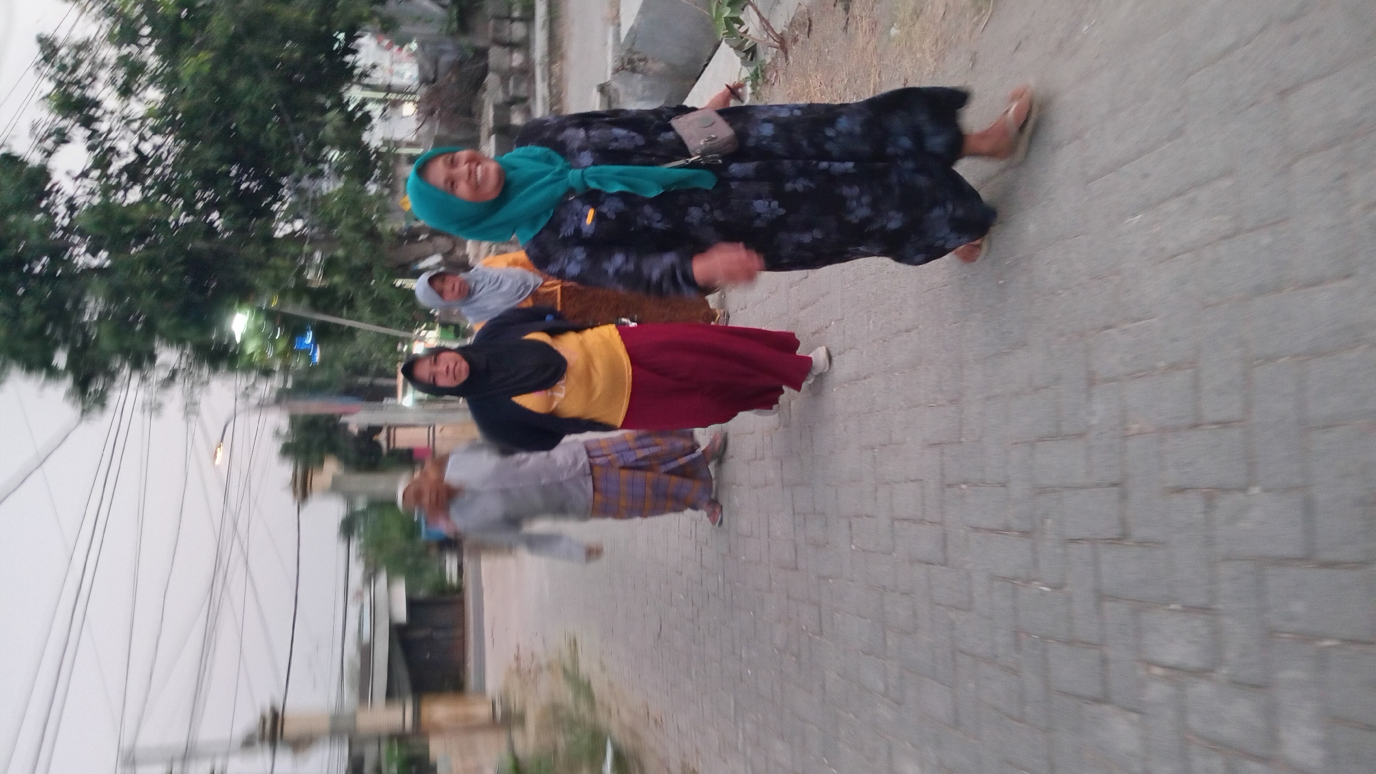 Four people are walking on a paved sidewalk in Trembulrejo, Indonesia. The first person, a man, is wearing a light-colored shirt and a sarong with a plaid pattern. The second person, a woman, is wearing a black hijab, a yellow shirt, and a red skirt. The third person, a woman, is wearing a light gray hijab and an orange shirt. The fourth person, a woman, is wearing a teal hijab and a dark blue dress with a floral pattern. She is smiling and carrying a small beige purse. The sidewalk is made of gray bricks. In the background, there are trees, buildings, and power lines. The sky is overcast.