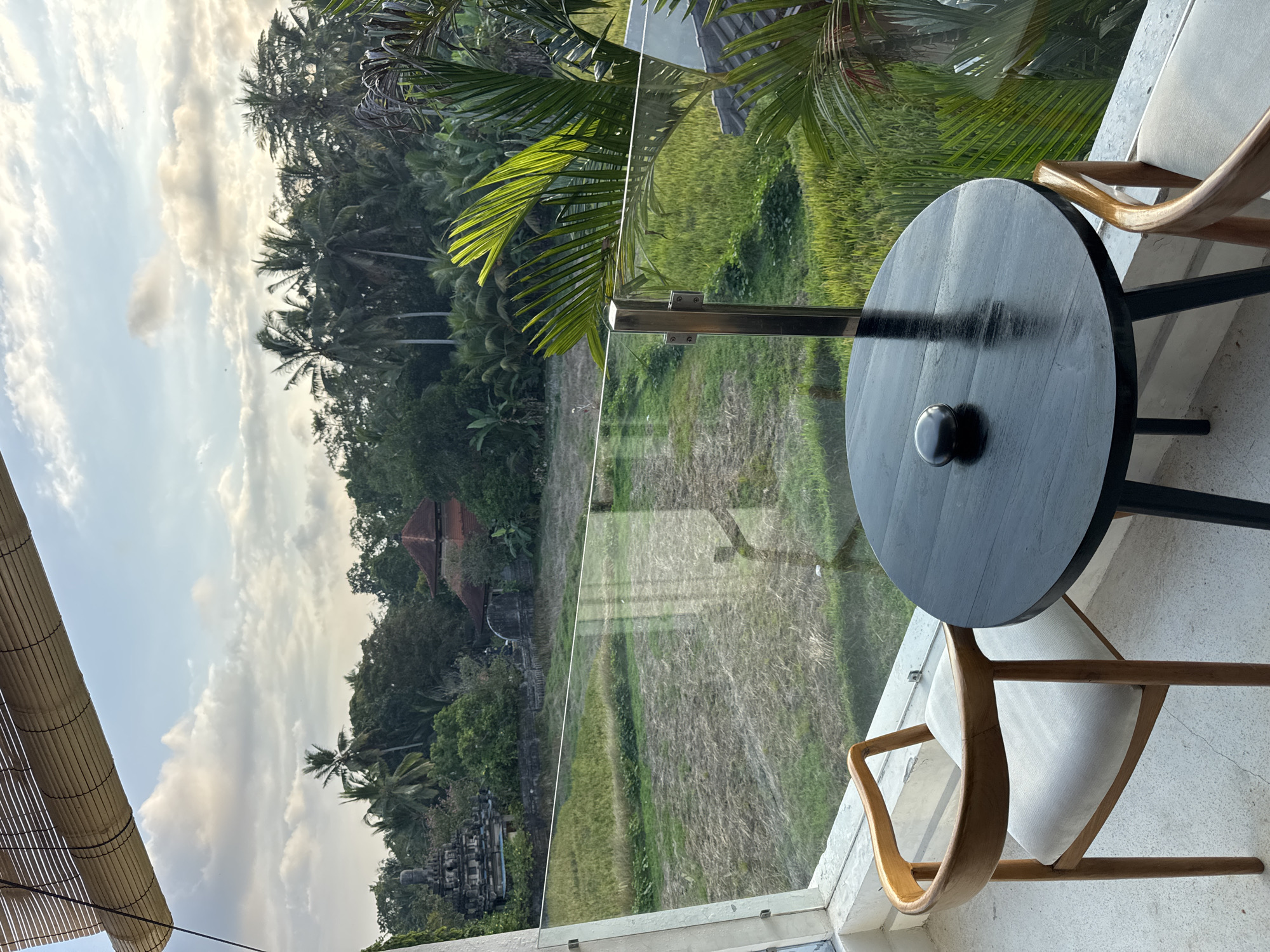 The image depicts a balcony in Tegallalang, Indonesia, offering a view of lush rice paddies and tropical vegetation under a partly cloudy sky. In the foreground, a round black table with a small dark object on it is positioned next to a wooden chair with a light-colored cushion. A glass railing separates the balcony from the scenic view. Behind the railing, green rice fields stretch out, bordered by a dense forest of palm trees and other tropical foliage. In the distance, traditional Indonesian buildings with terracotta roofs are visible amidst the greenery, along with what appears to be a stone temple structure. The lighting suggests it is either early morning or late afternoon.