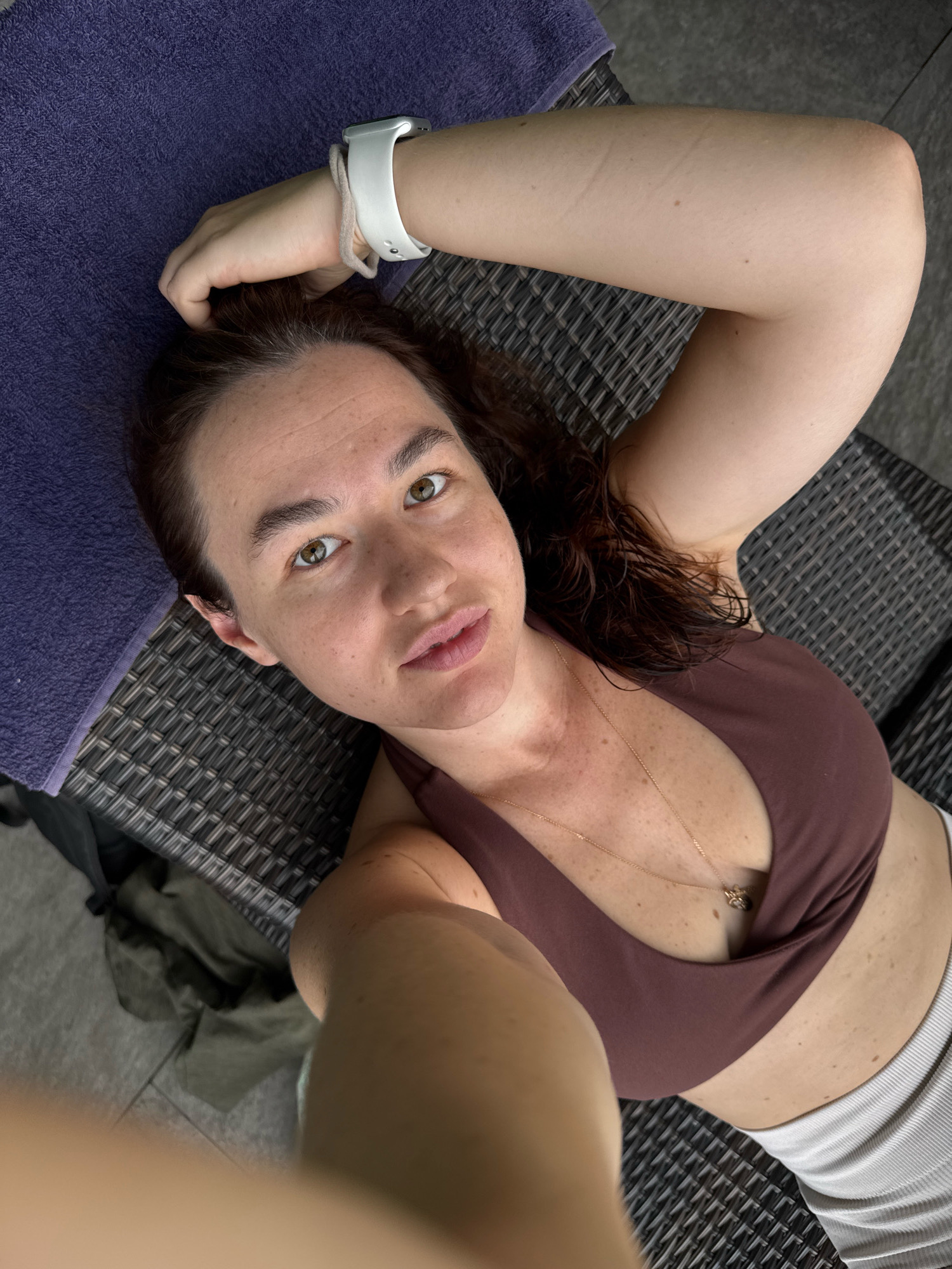 A woman with brown hair and brown eyes is lying on a lounge chair outdoors. She is wearing a brown bikini top and white shorts. A purple towel is draped over the back of the lounge chair. The background is blurred, but appears to be a tiled patio. The image was taken in Samrong Nuea Subdistrict Municipality, Thailand.