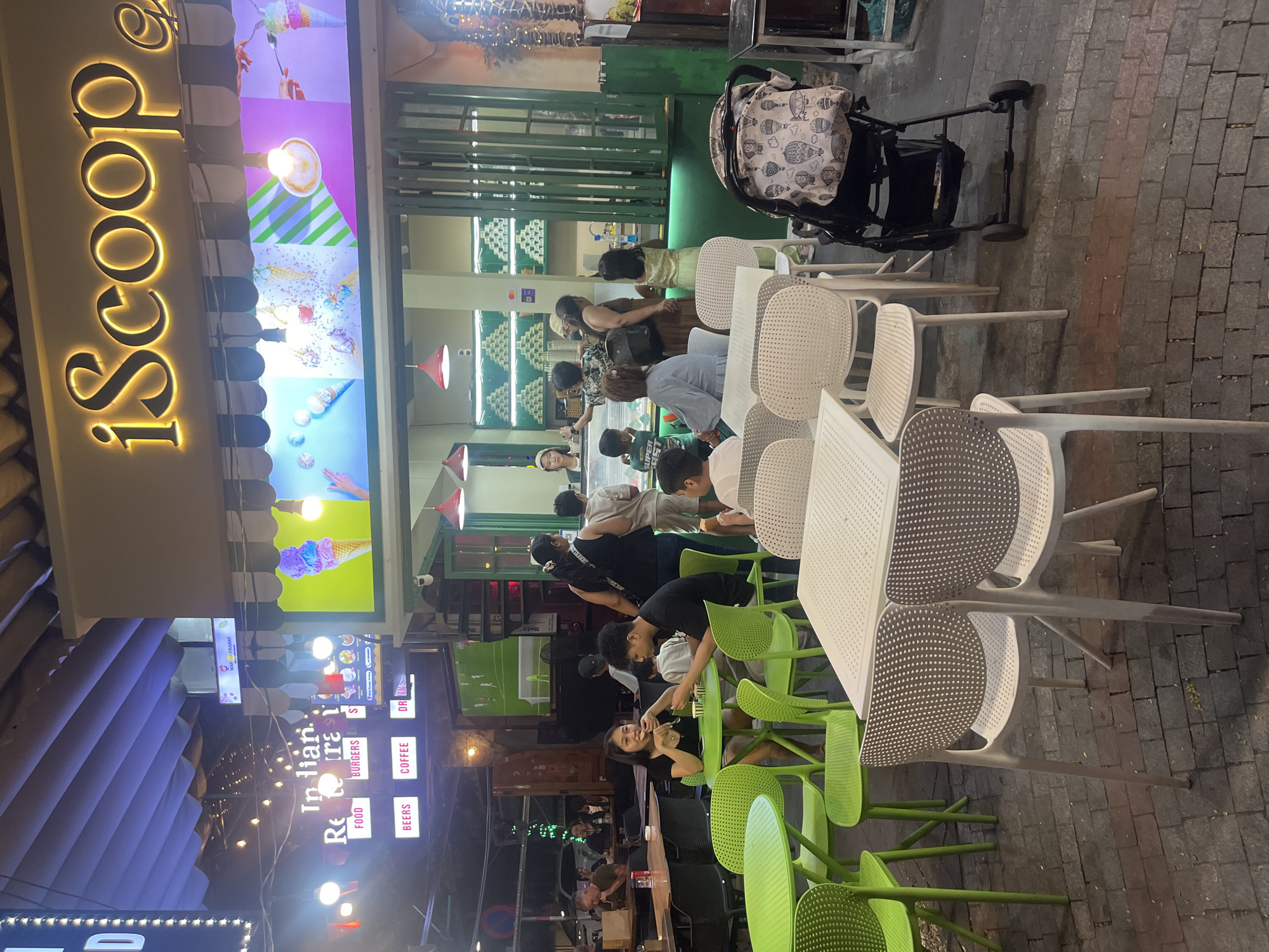 The image captures an outdoor dining area at "iScoop" ice cream shop in Đà Nẵng, Vietnam. The storefront features a prominent "iScoop" sign with illuminated lettering, above a colorful display of ice cream images. In front of the shop, several green plastic tables and chairs are set up on a cobblestone pavement. A couple, a woman and a man, are seated at one of the green tables, eating ice cream. Other patrons are visible in the background, including children and adults near the counter of the ice cream shop. A black stroller with a patterned bag is parked to the right of the dining area. The scene is illuminated by ambient lighting, with a visible television screen showing a soccer match in the background. The general atmosphere suggests a casual evening gathering at a popular dessert spot.
