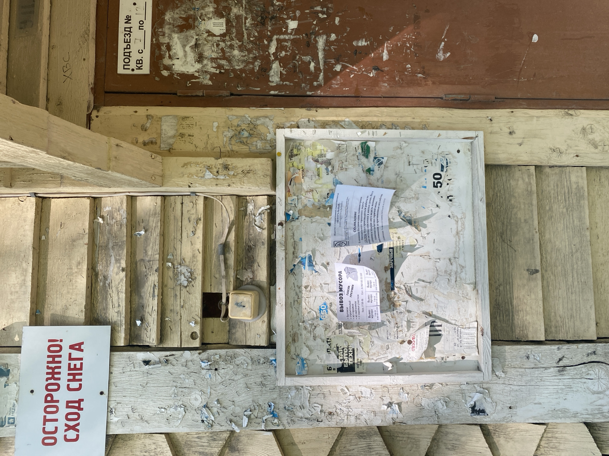 This is an outdoor shot of a weathered, cluttered bulletin board and surrounding structure, likely in Novouralsk, Russia, given the Cyrillic signage. The bulletin board, framed in white, is covered with torn notices and scraps of paper. Amongst the scattered ads, there's a notice for "Vyvoz Musora" (Garbage Removal), and an announcement mentioning the "Centralny" (Central) company and temporary disruptions in hot water quality. To the left of the board is a red and white sign warning "Ostorozhno! Skhod Snega" (Caution! Snow Coming Down). An old-fashioned electrical outlet is also visible. The wood of the structure is peeling and distressed, with numerous staples left from previous postings. The lighting suggests it is daytime and fairly bright.