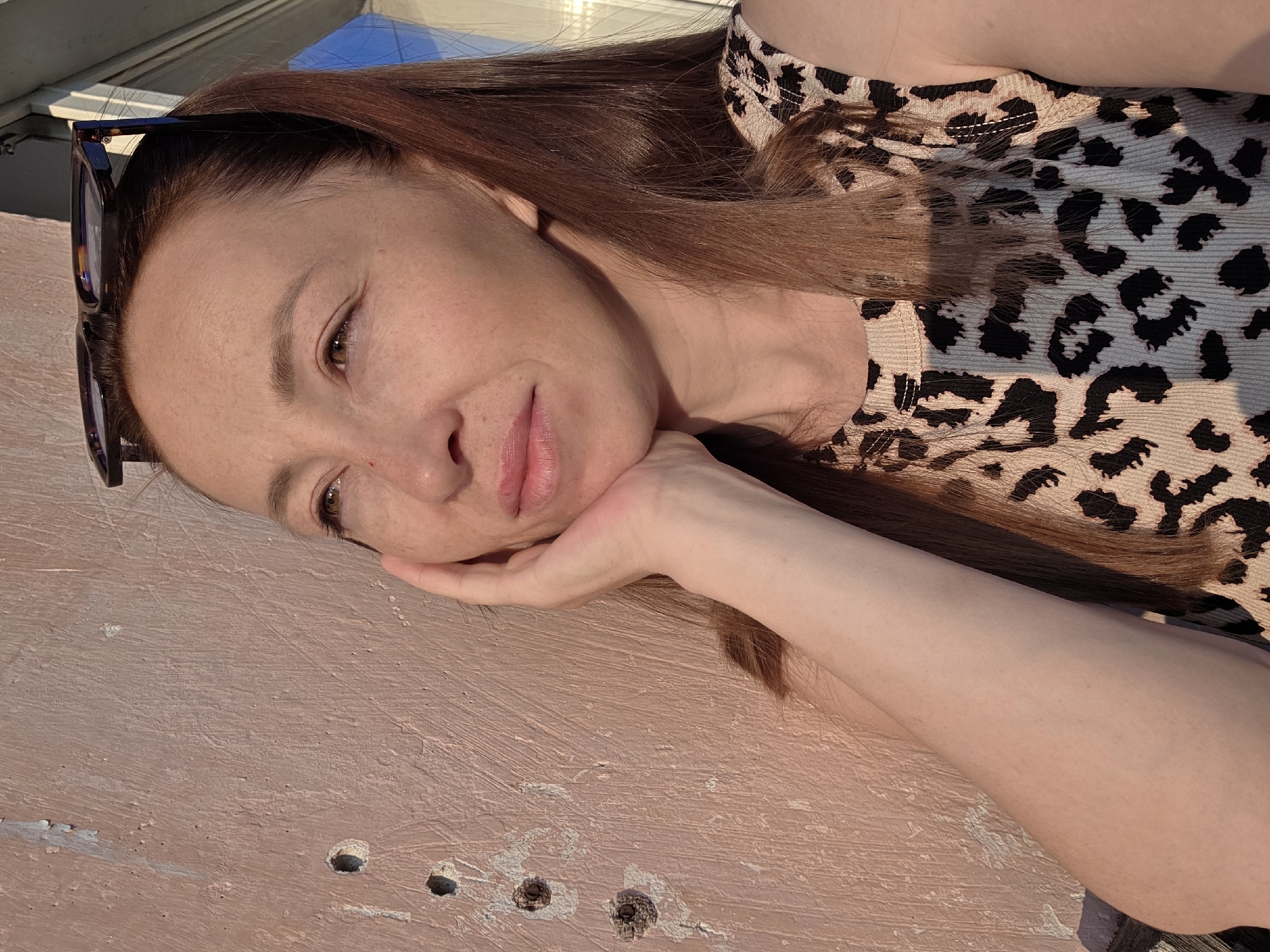 A medium-close shot captures a woman with auburn hair, wearing a sleeveless leopard-print top. She is resting her chin on her left hand, with her eyes looking upward and to the left, bathed in the warm light of what appears to be sunset. She has freckles and is wearing eyeglasses pushed up on her head. She is positioned against a textured, pinkish wall with several holes and some peeling paint, possibly a weathered exterior surface. The background includes a bright, out-of-focus area that suggests an outdoor setting, possibly near a window or building. The lighting indicates a time of day with low sun angle, casting soft shadows.